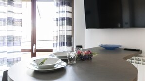 Ein Essbereich mit einem runden Tisch, Tellern, einem Glas und einer Schüssel. Ein Flachbildfernseher ist an der Wand befestigt.
