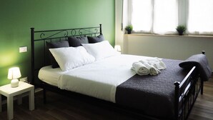 Una cama bien hecha con almohadas blancas y oscuras, una toalla doblada y una manta oscura. Junto a la cama hay una mesita con una lámpara. La habitación tiene paredes verdes y una ventana con persianas.