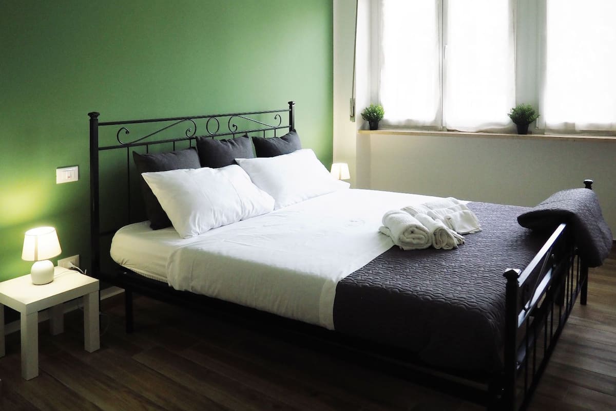 A neatly made bed with white and dark pillows, a folded towel, and a dark blanket. There is a small table with a lamp beside the bed. The room has green walls and a window with blinds.
