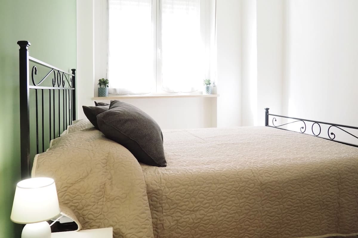 A bedroom with a bed, a window with curtains, a bedside table with a lamp, and a small plant on the windowsill.