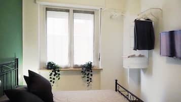 A bedroom with a bed, a window with curtains, a TV mounted on the wall, and a small shelf with a plant.