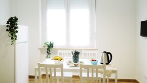 Una cocina pequeña y luminosa con paredes blancas, una ventana con persianas, un refrigerador blanco, una mesa blanca con sillas y una tetera sobre la mesa.