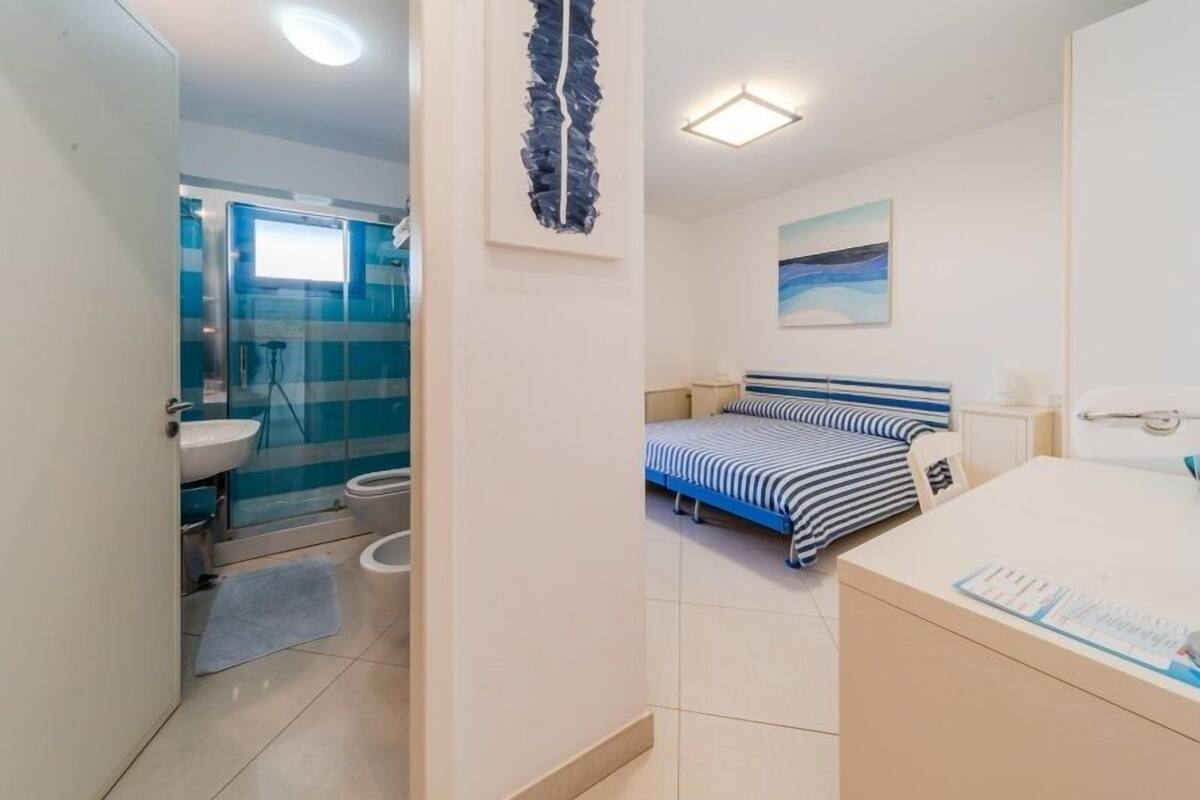 A bedroom with a bed, a bedside table, a window, and a bathroom with a sink and toilet.