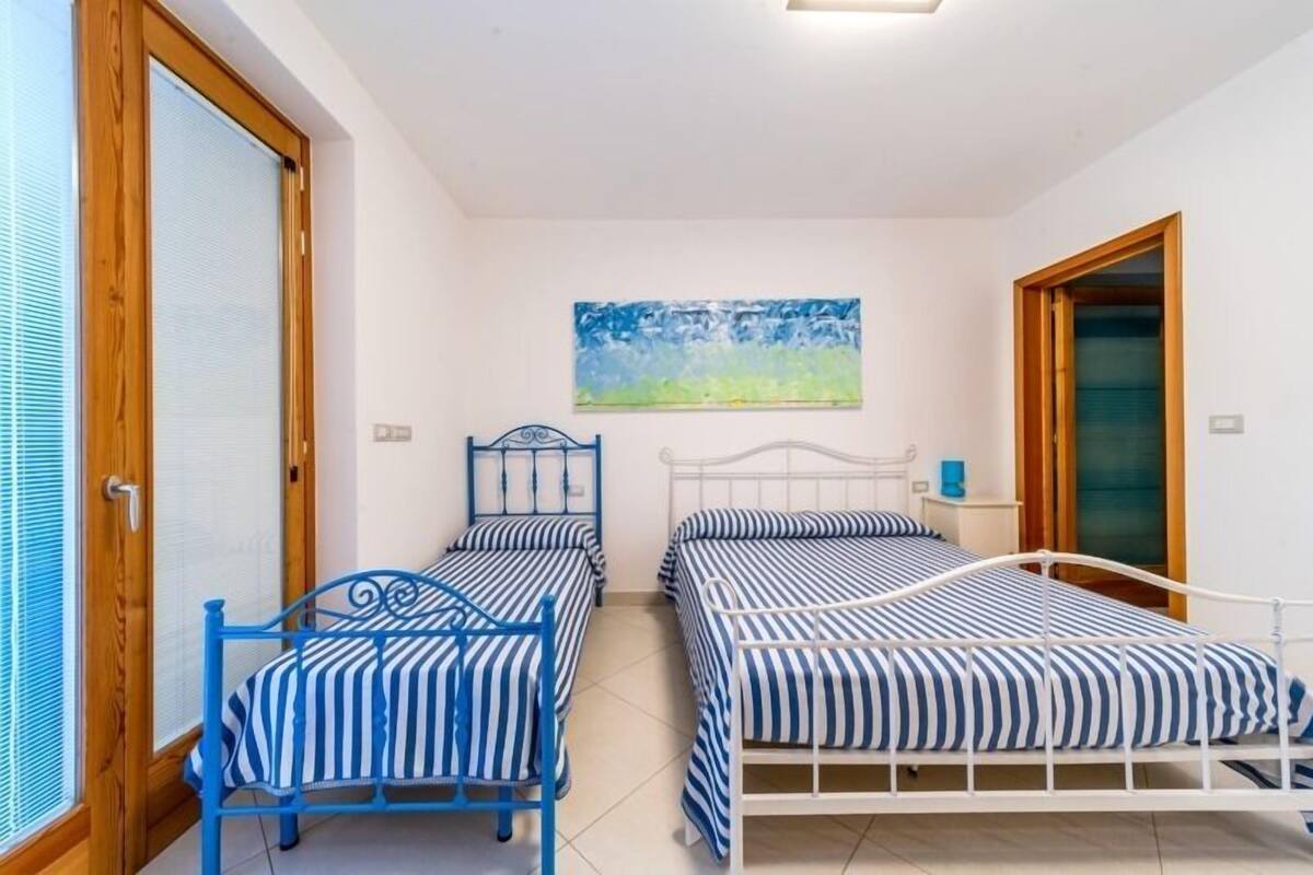 A bedroom with two beds, a painting on the wall, and a window with blinds.