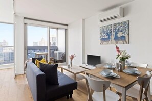 A modern living room with a sofa, dining table, and chairs. There is a TV on a stand, a vase with flowers, and a painting on the wall.
