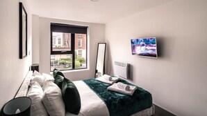 A modern hotel room with a large bed, a flat-screen TV, a window with a city view, and a standing lamp. - Cozy 1-BR | Steps from Liverpool Centre (Liverpool)