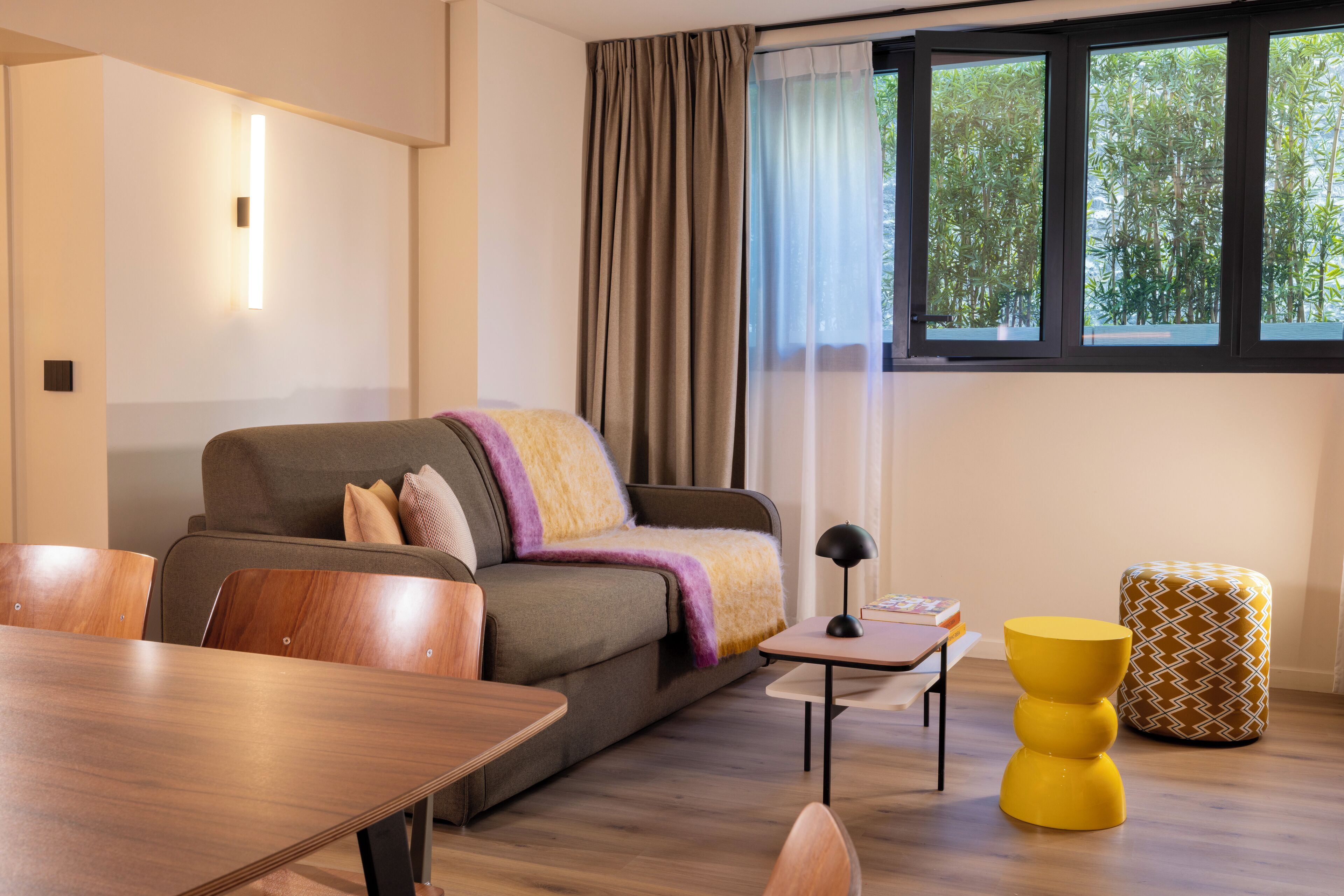 A modern living room with a sofa, wooden chairs, a small table, and a yellow ottoman.