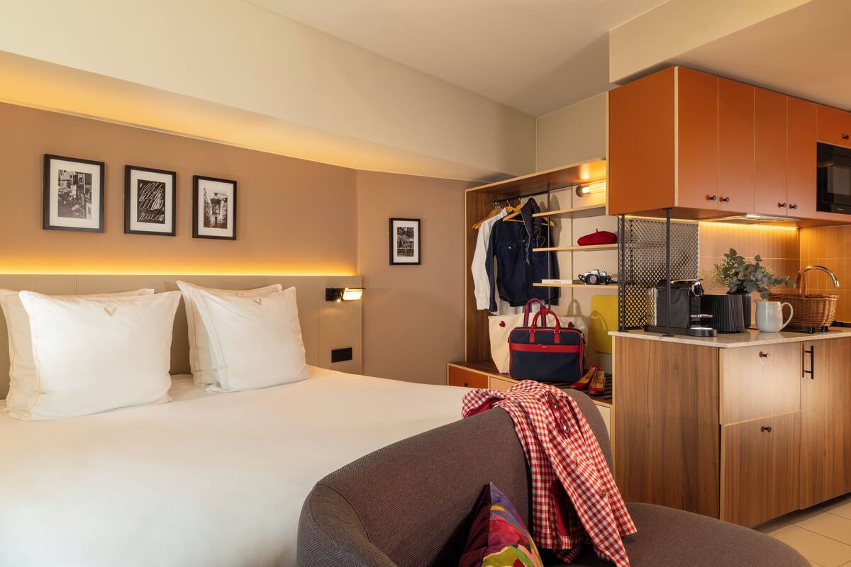 A hotel room with a bed, a sofa, a kitchenette, and framed pictures on the wall.