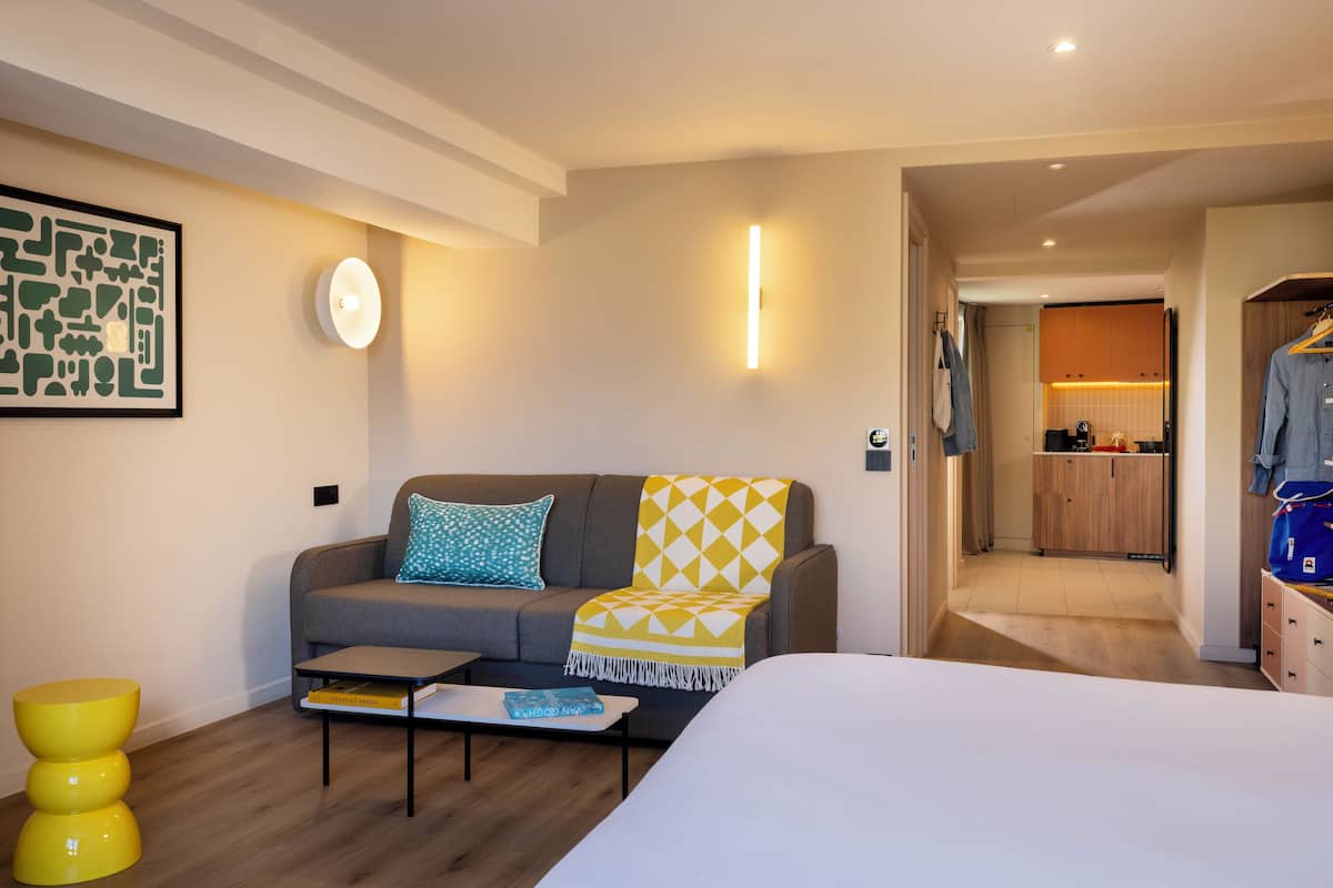 A modern hotel room with a sofa, a coffee table, a bed, and a kitchen area in the background.