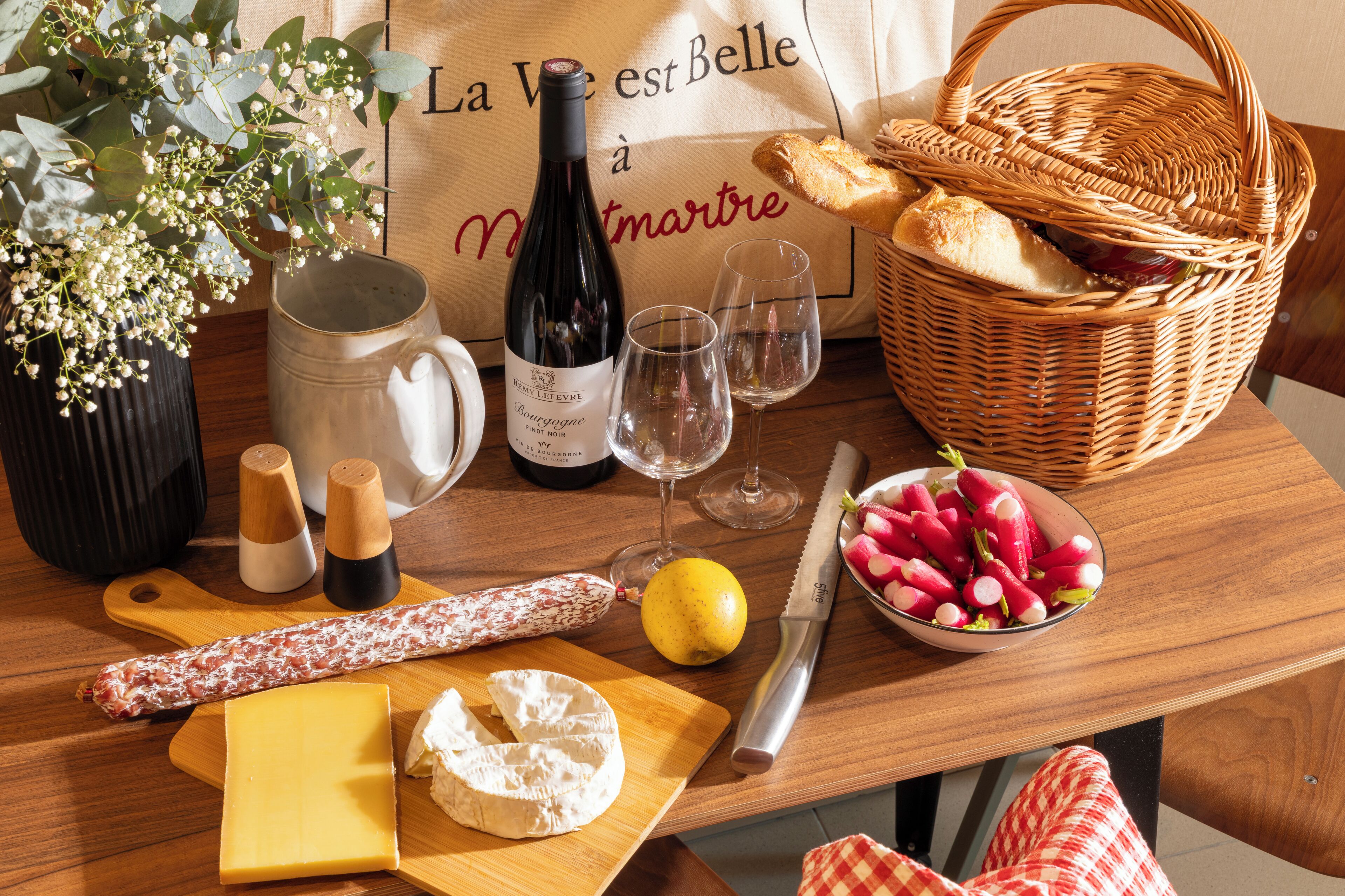 A wooden table with a bottle of wine, glasses, a knife, a bowl of radishes, a loaf of bread, a cheese wheel, and a baguette.