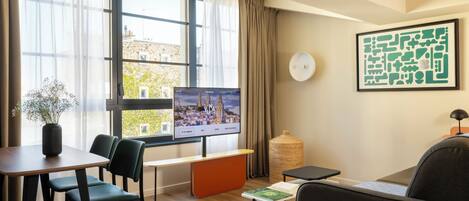 Una habitación de hotel moderna con un ventanal grande, un televisor de pantalla plana, un área de comedor con mesa y sillas, un sofá, una mesita auxiliar pequeña y un jarrón decorativo.
