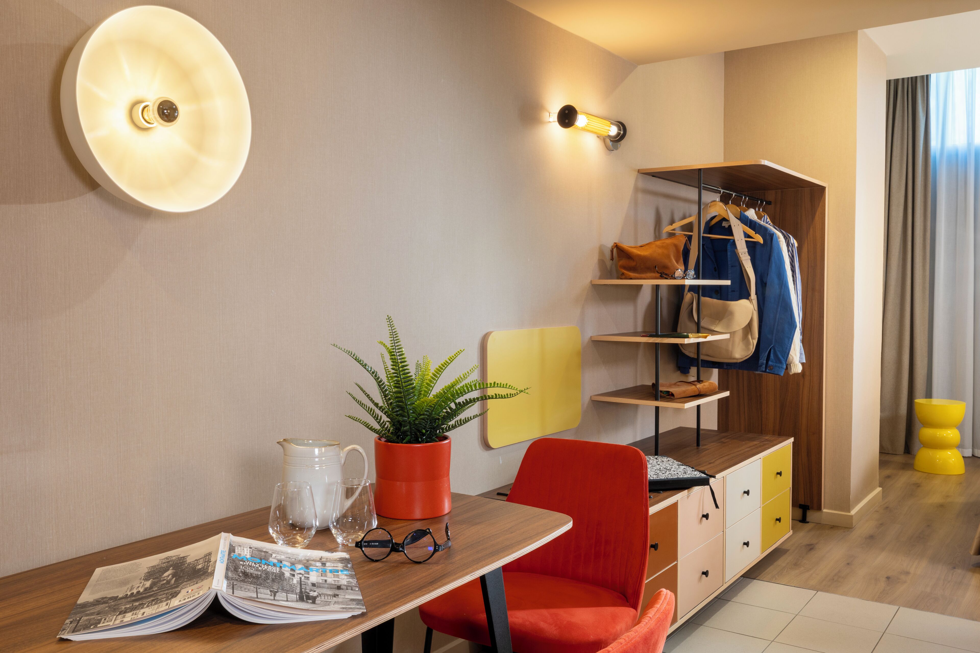 Une chambre d’hôtel moderne avec un bureau en bois, une chaise rouge, une petite plante, un porte-manteau et des toilettes jaunes.