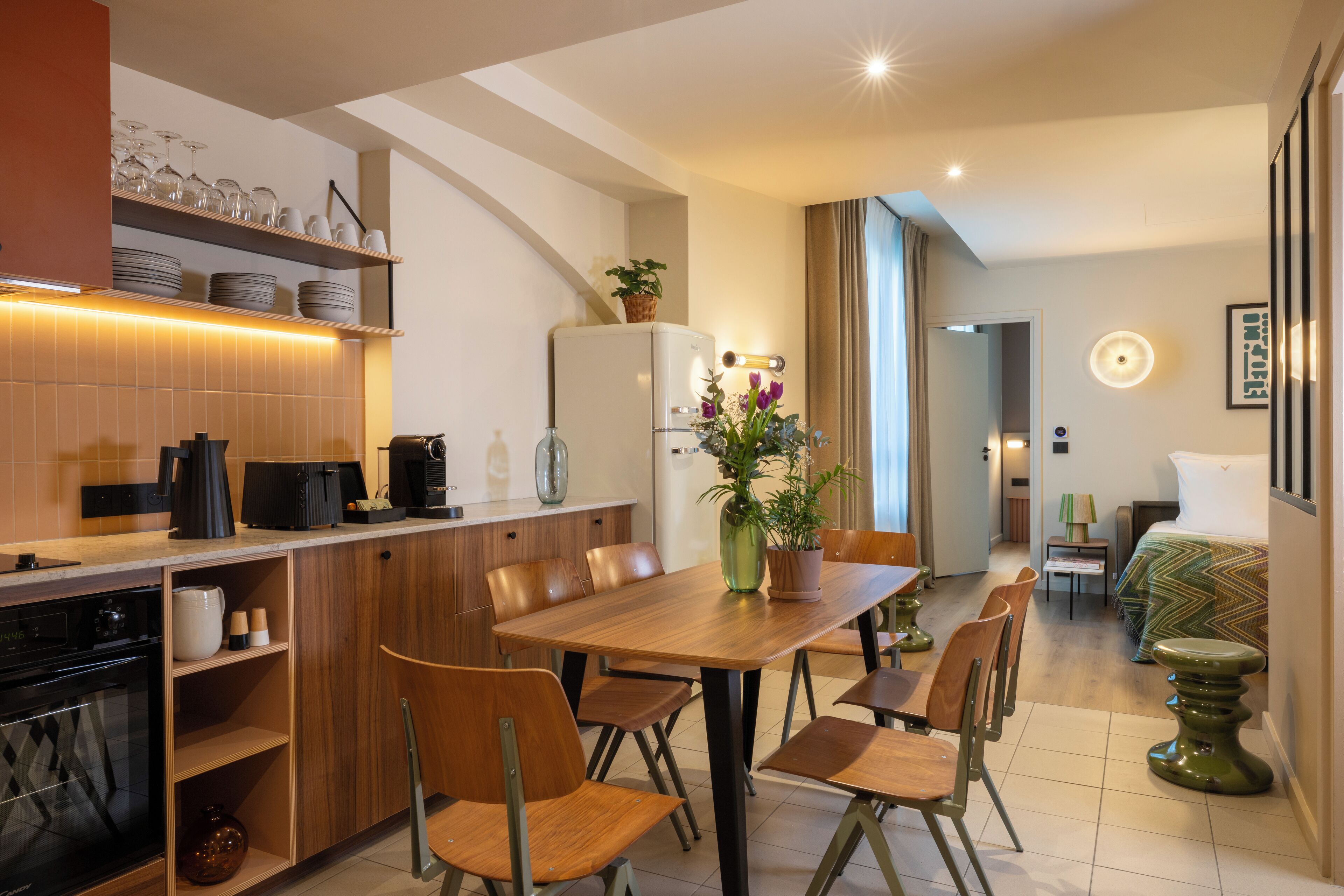 A modern kitchen with wooden cabinets, a dining area with a wooden table and chairs, a refrigerator, and a small dining table with a lamp.