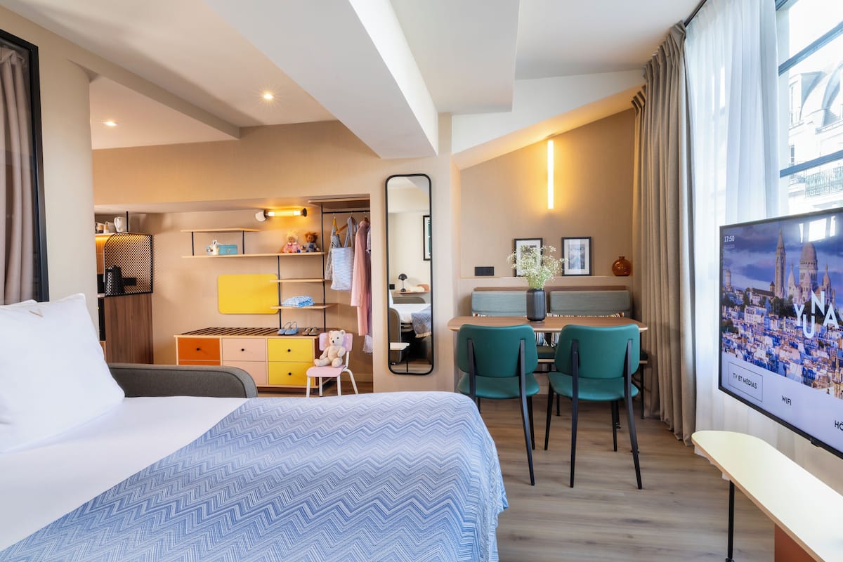 A hotel room with a bed, a desk, chairs, a TV, and a view of a cityscape.