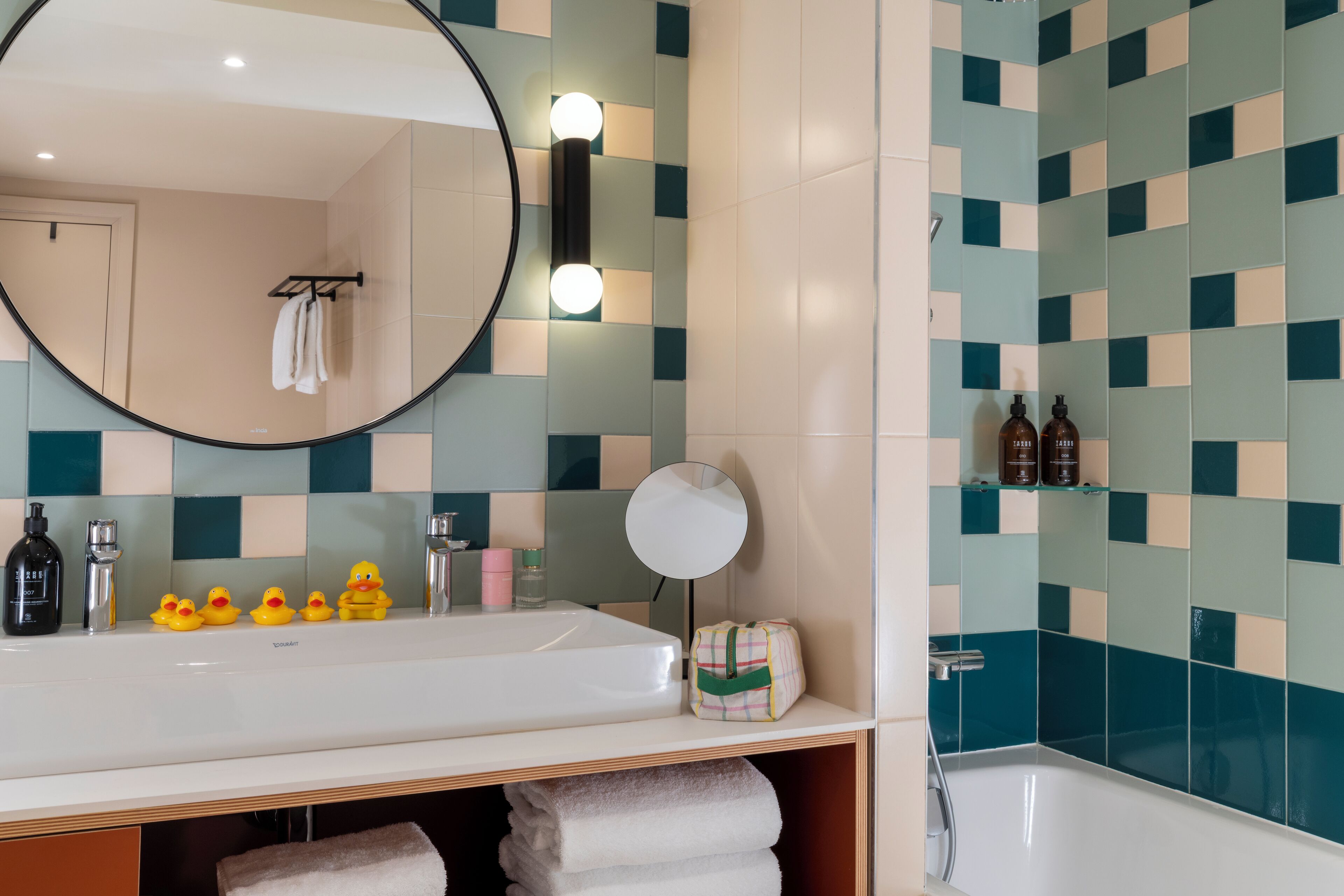 A bathroom with a large round mirror, a sink, and a bathtub. There are toiletries on the sink and a towel rack above it.