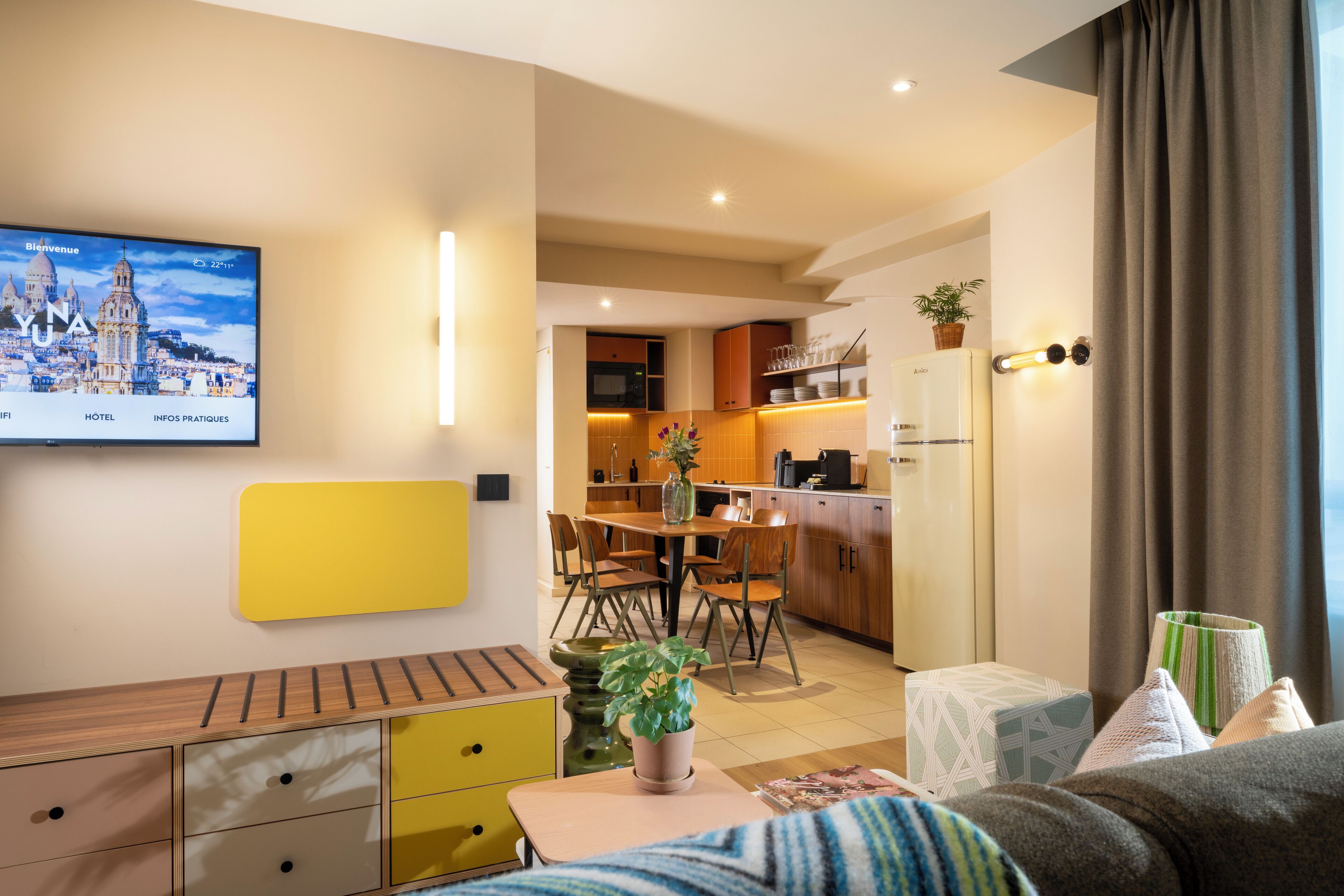 A modern hotel room with a dining area, kitchenette, and a TV displaying a cityscape.