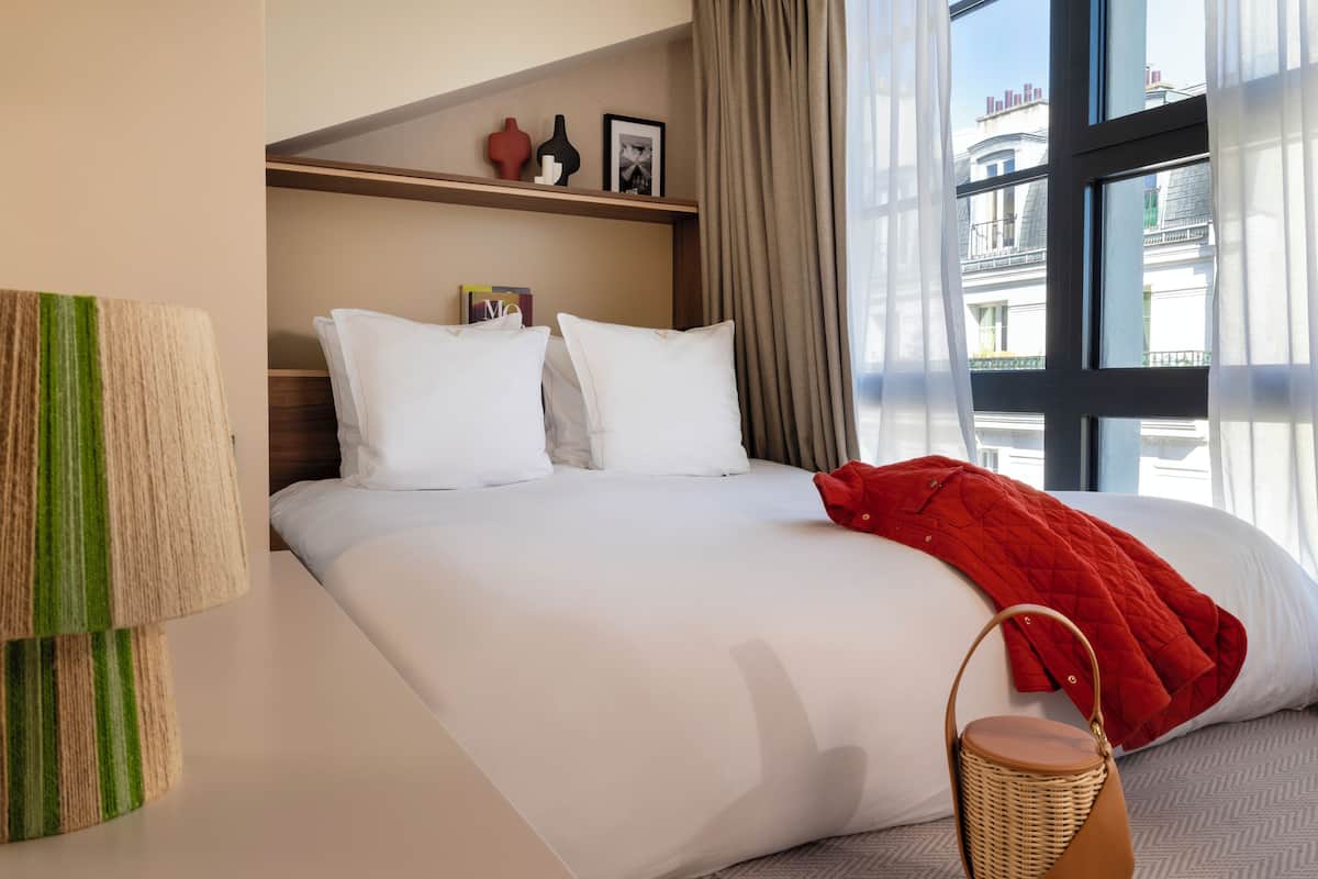 A hotel room with a large bed, a red blanket, a wicker basket, and a view of buildings through the window.