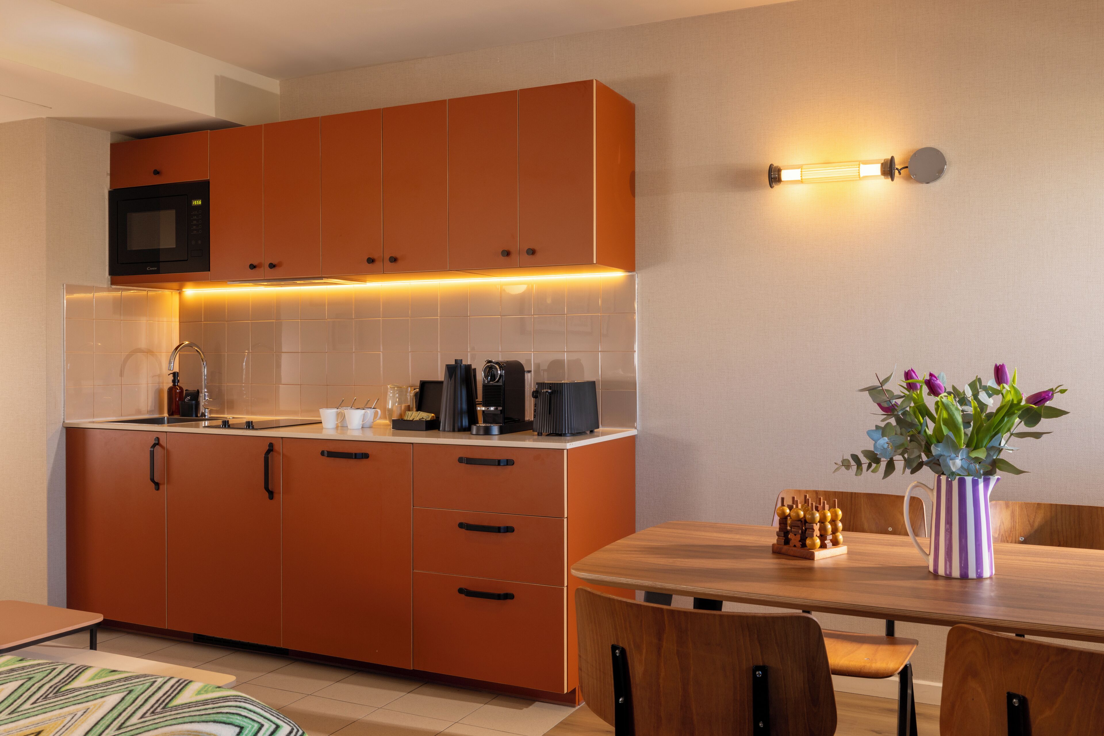 A modern kitchen with orange cabinets, a microwave, and a coffee machine. There is a dining table with chairs and a vase of flowers.