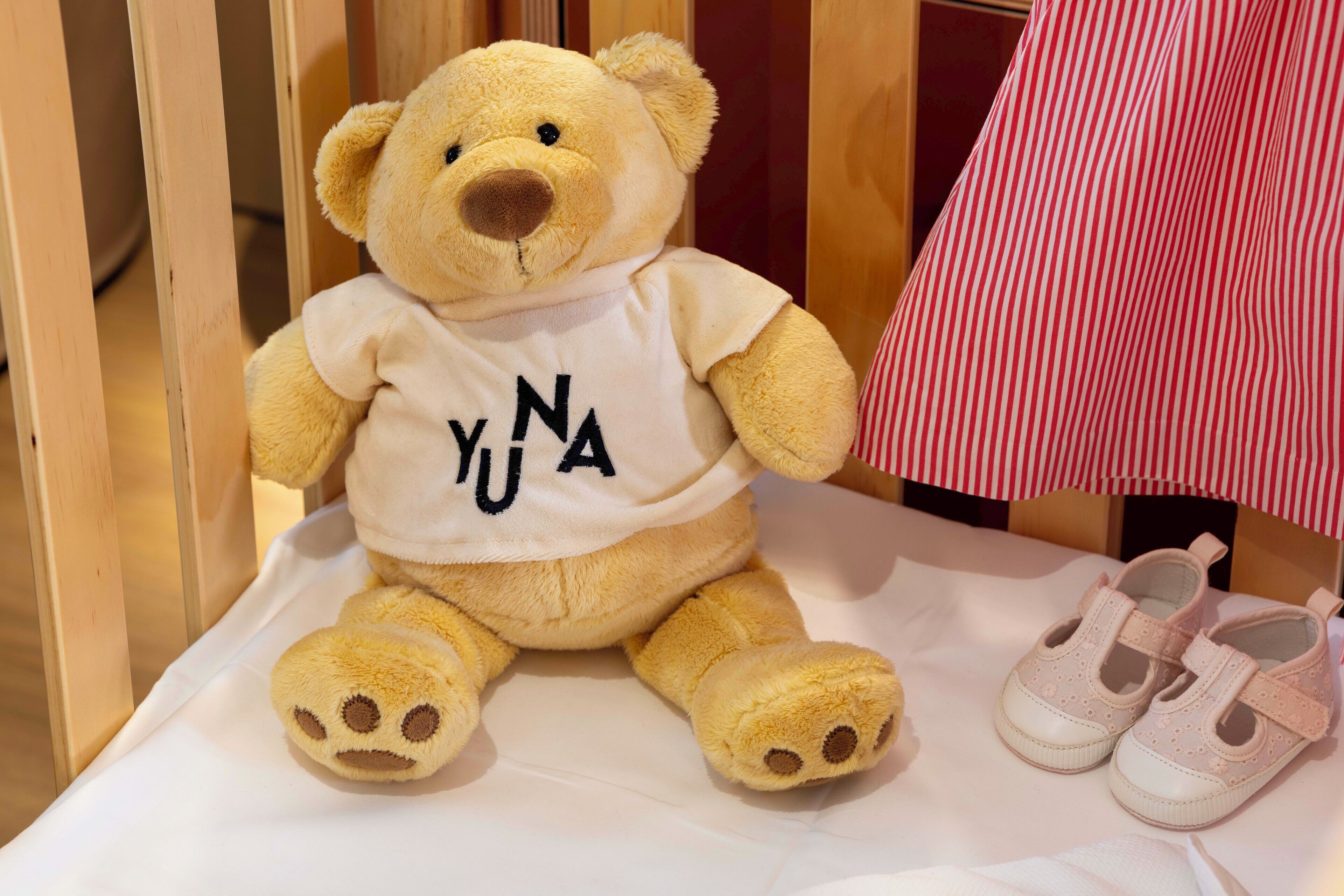 A plush teddy bear with a white shirt and a pair of baby shoes on a white surface.