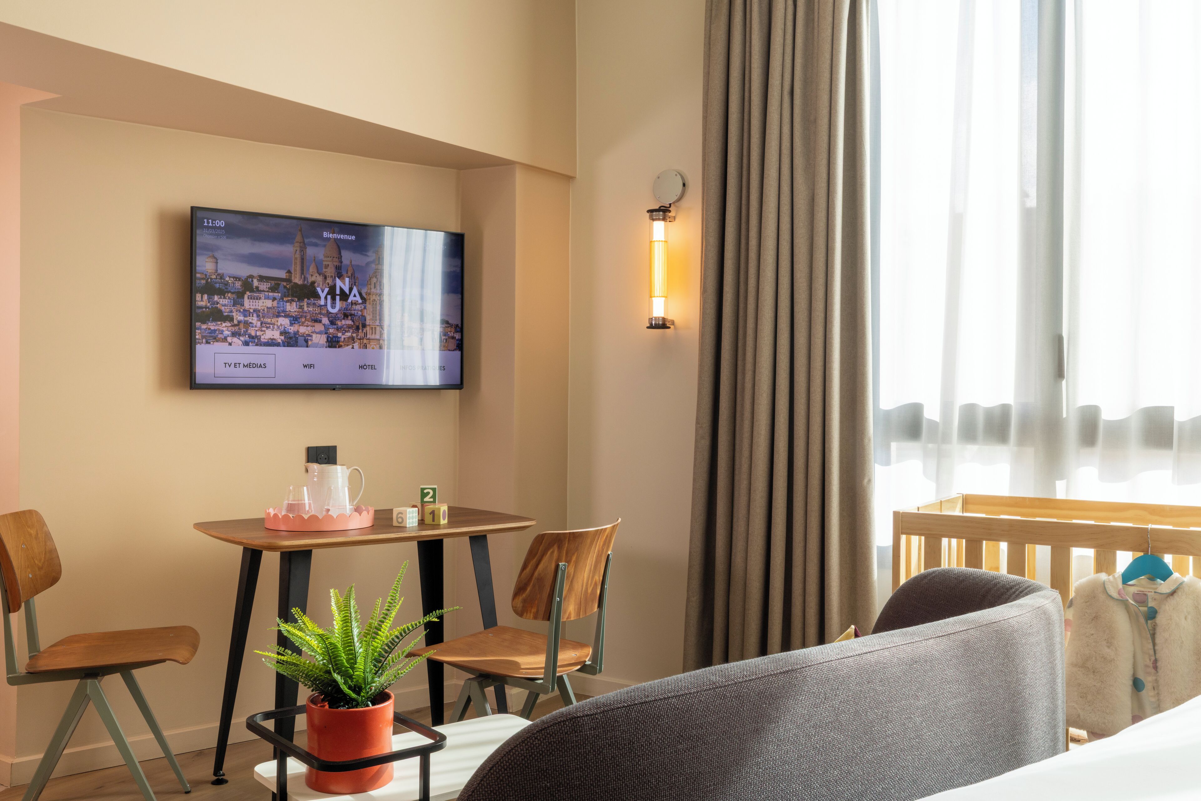 Ein Hotelzimmer mit Fernseher, der eine Stadtansicht zeigt, einem kleinen Tisch mit einer Pflanze, zwei Stühlen und einem Bett mit aufgehängtem Mantel.