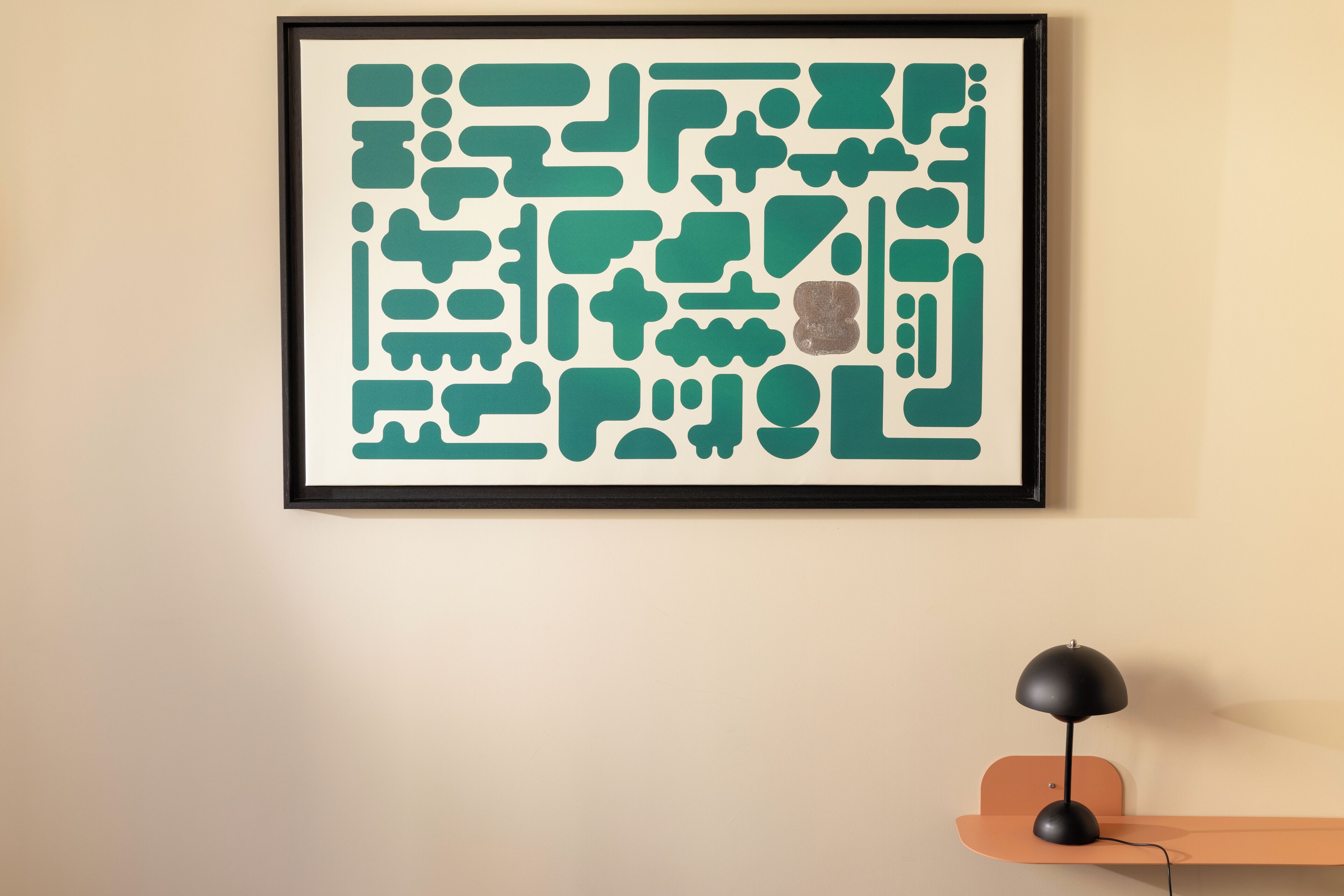A framed abstract art piece on a wall, a black desk lamp on a wooden stand, and a plain wall.