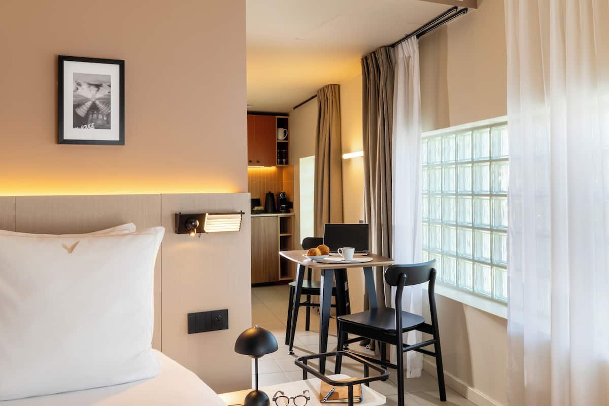 A hotel room with a bed, a desk with a chair, a window with curtains, a framed picture on the wall, and a kitchenette in the background.