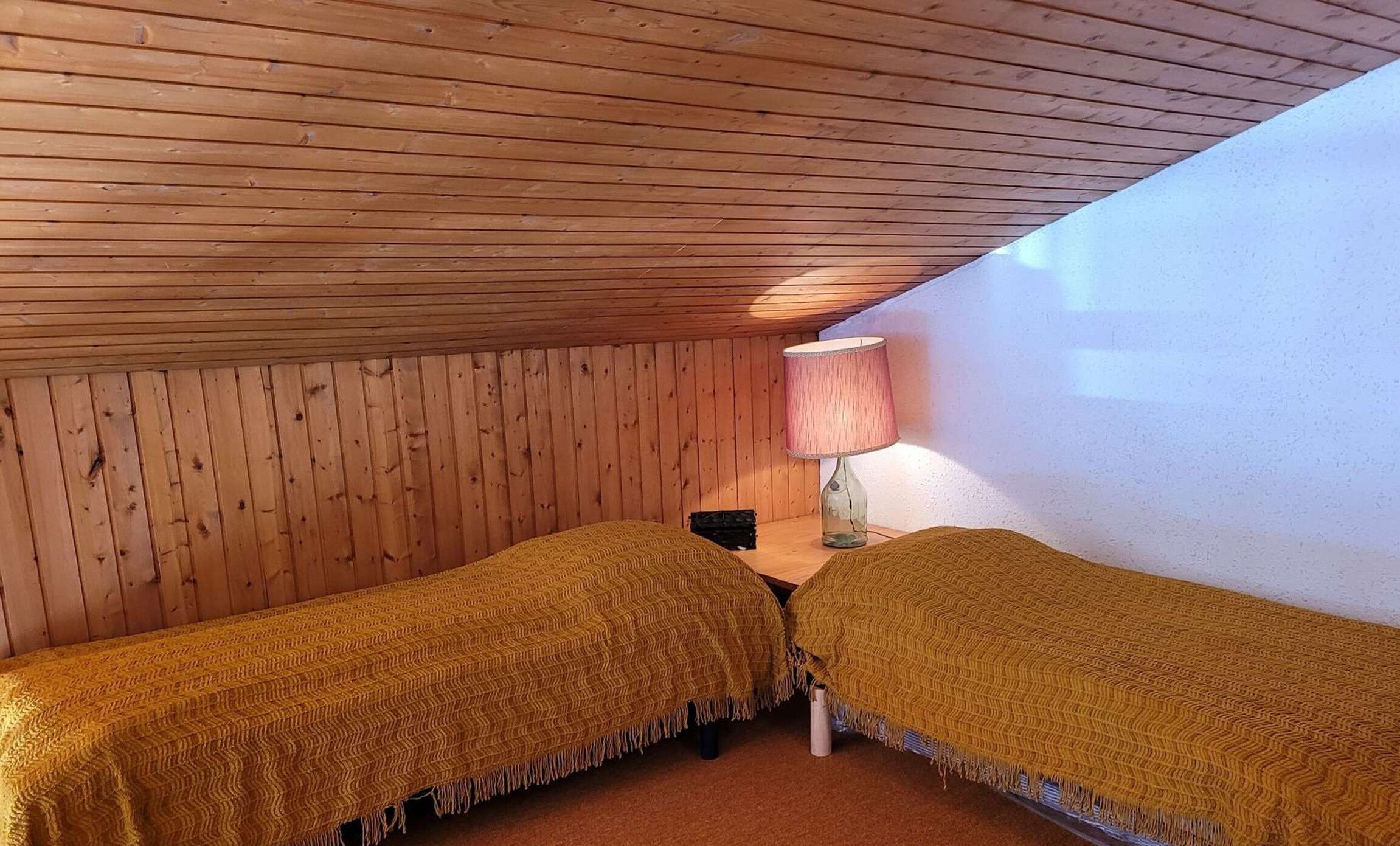 A small, cozy room with wooden walls and a sloped ceiling, featuring two beds with matching blankets and a lamp on a nightstand.