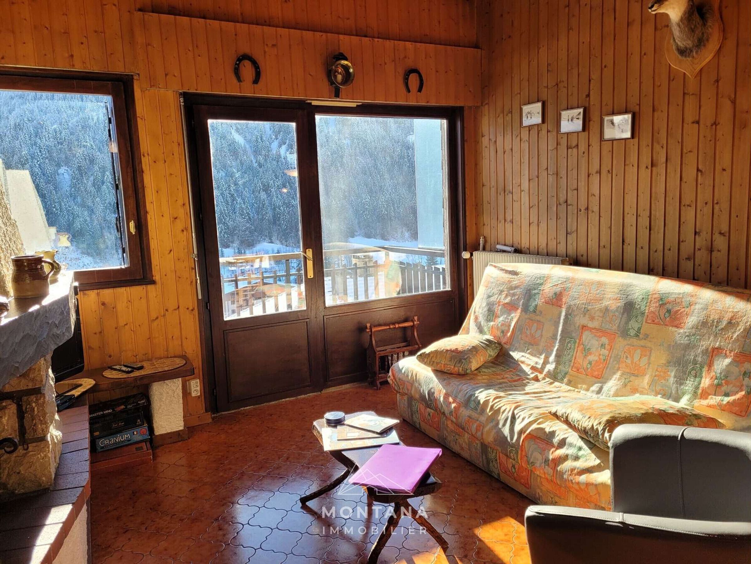 Ein gemütliches Blockhaus-Interieur mit Holzausstattung, einer Couch, einem Tisch mit einem Laptop und Blick auf eine verschneite Landschaft durch große Fenster.