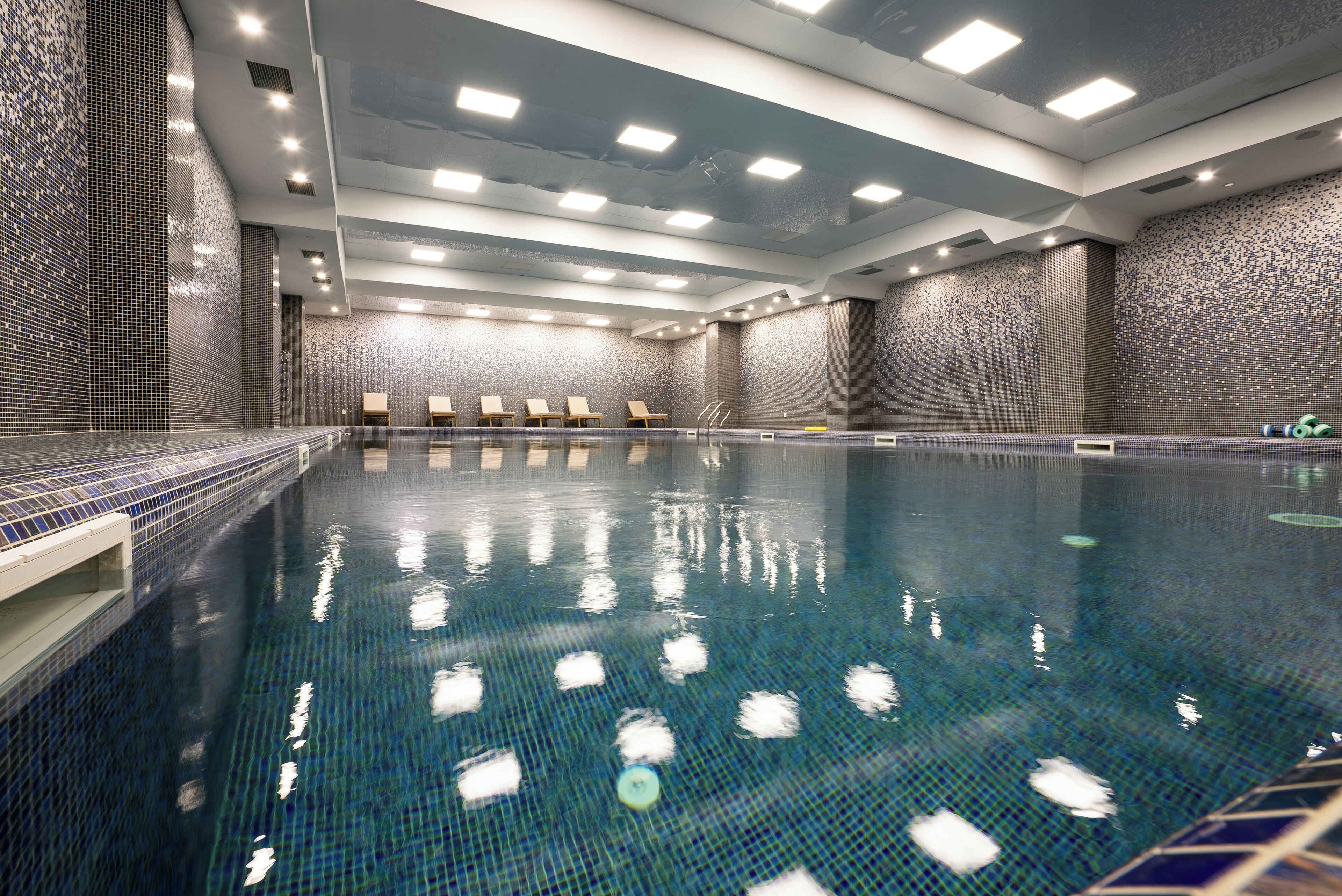 Una piscina con paredes de azulejos, luces en el techo y sillones de descanso.
