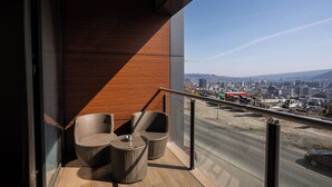 Ein Balkon mit Korbstühlen und einem kleinen Tisch, mit Blick auf eine Stadtlandschaft mit Gebäuden und einer Straße.