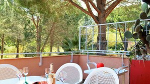 An outdoor dining area with a table set for two, surrounded by white chairs, a red wall, and a metal railing overlooking a garden with trees and cacti. - Charming 2-Bedroom Apartment with Pool and Terrace in Le Lavandou for 6 (Le Lavandou)