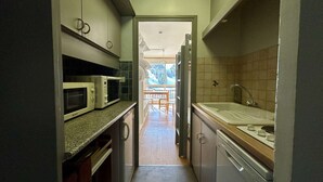 A narrow kitchen with a microwave, oven, and sink. There is a wooden table and chairs in the background.