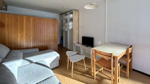 A modern, compact room with a wooden wardrobe, a sofa, a small table with chairs, and a TV.