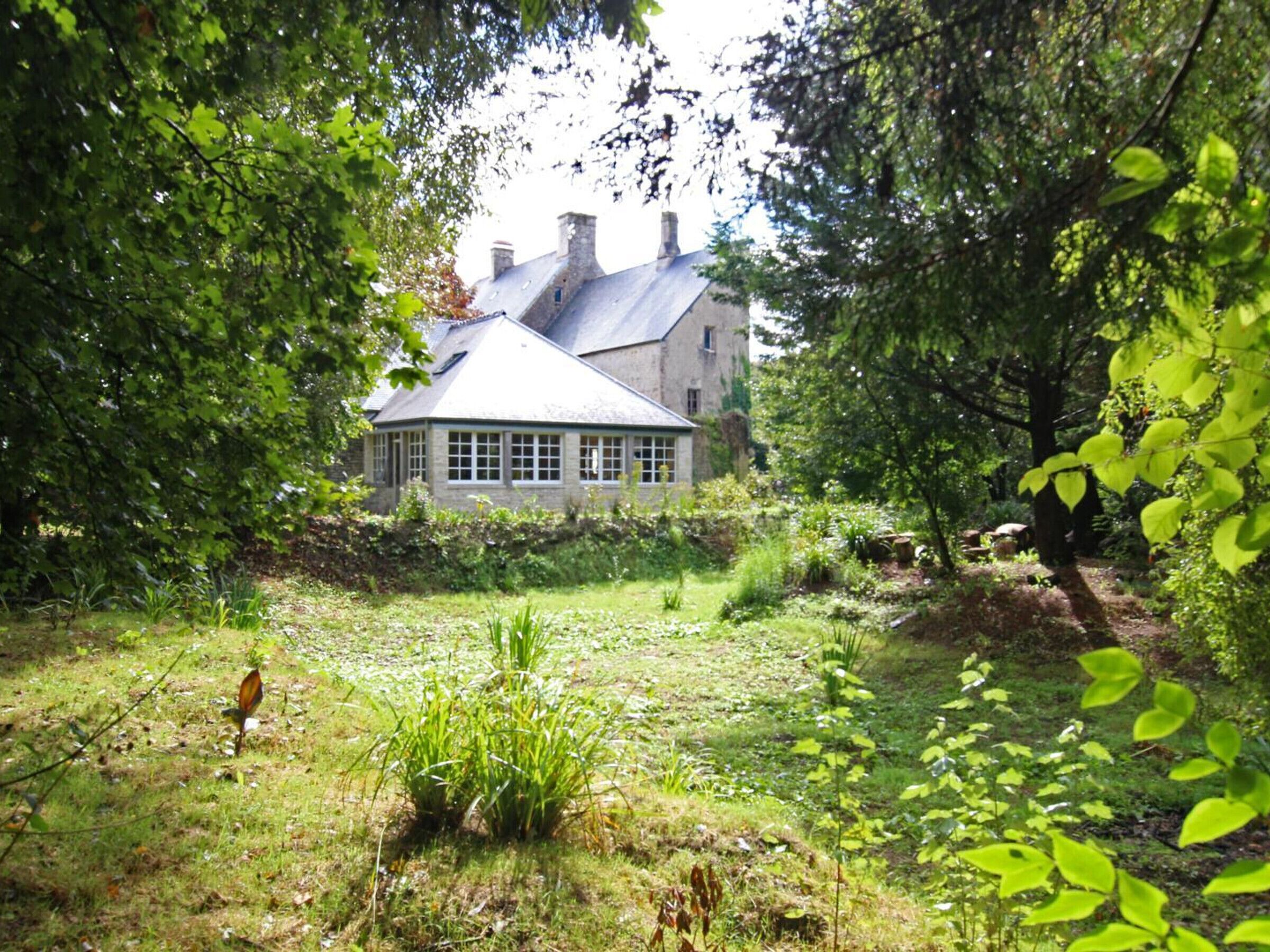 Une maison de campagne en pierre, dotée d’un toit à pignon, entourée d’une végétation luxuriante et d’un jardin bien entretenu.