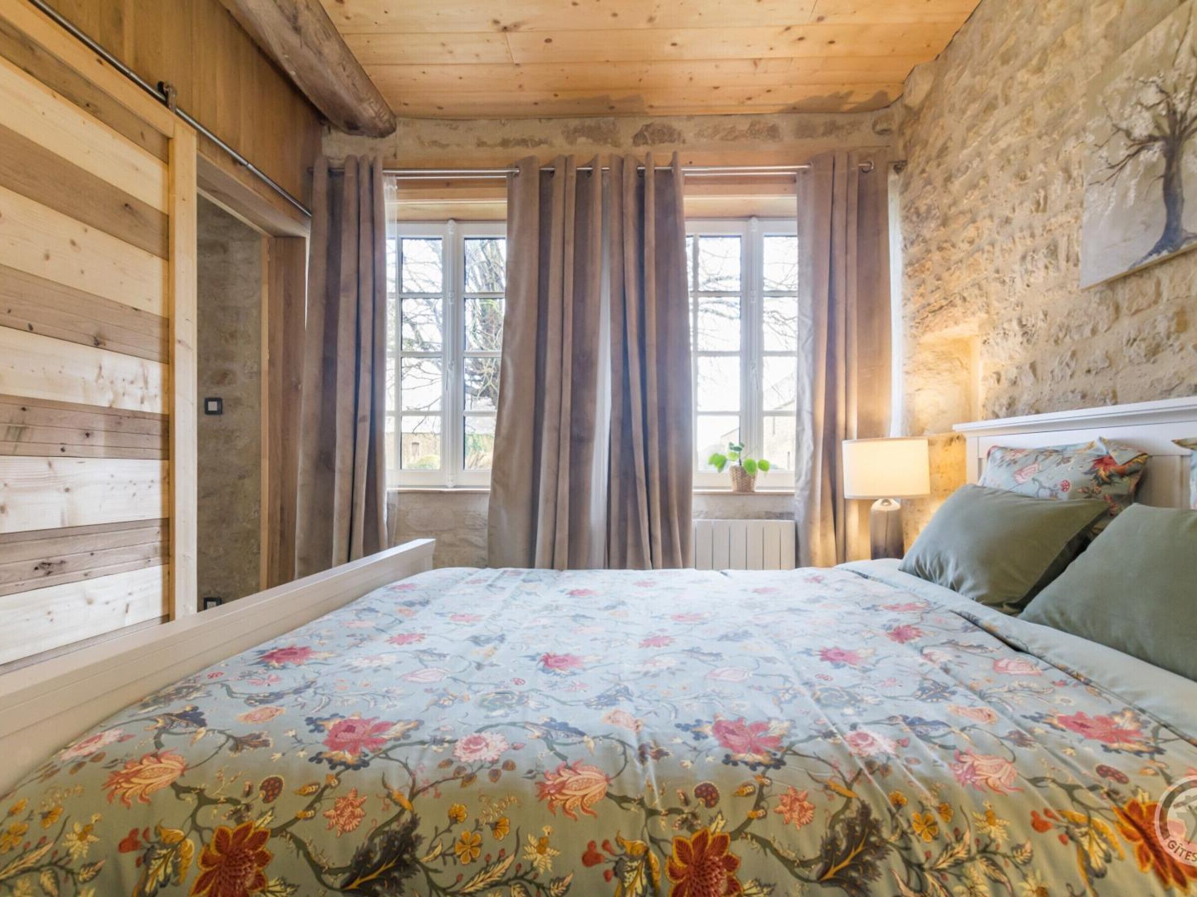 A bedroom with a wooden ceiling, a bed with floral bedding, a window with curtains, and a stone wall.