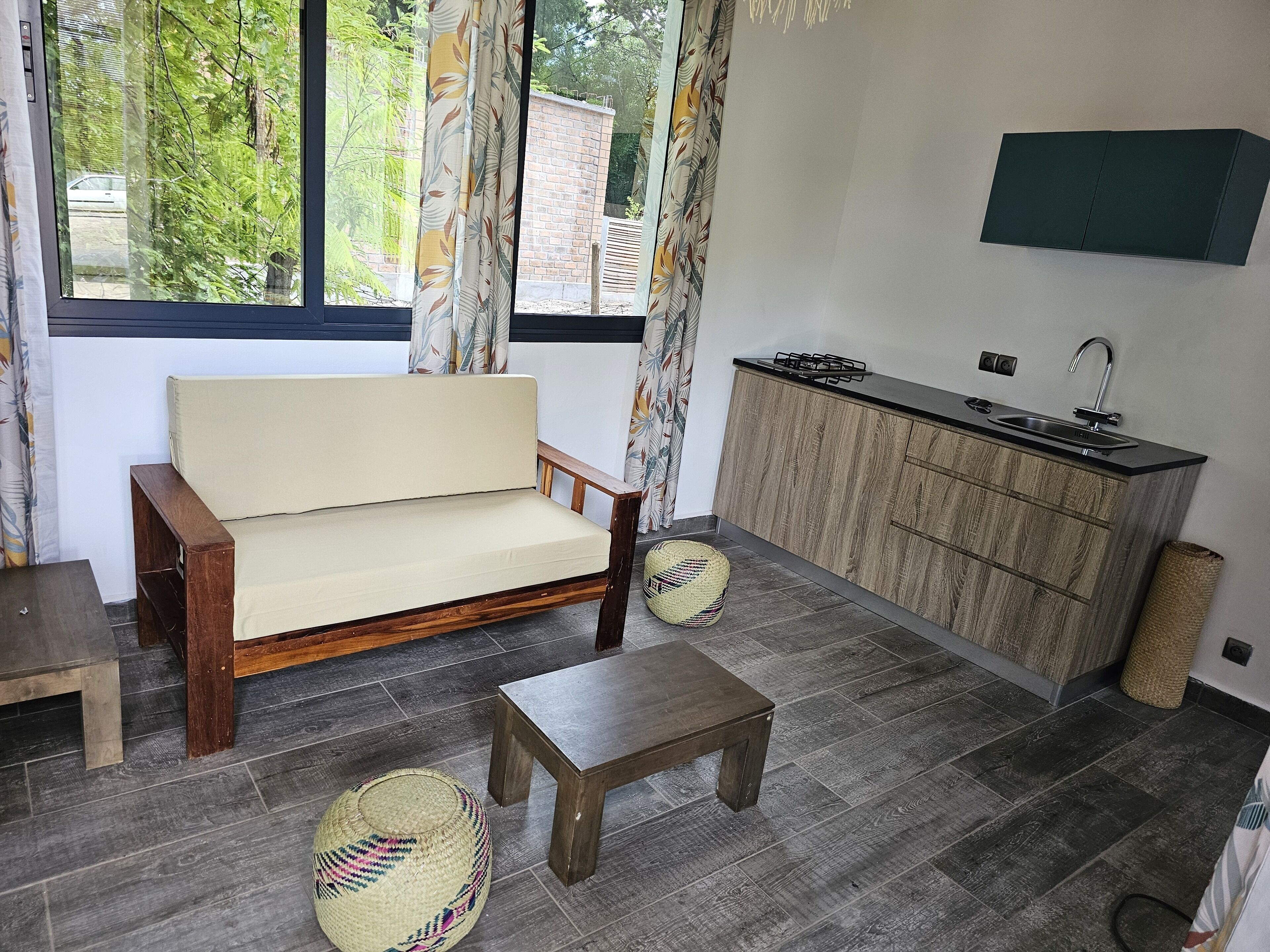 A compact living space with a sofa, a small table, a woven pouf, a kitchen area with a sink and stove, and a large window with curtains.