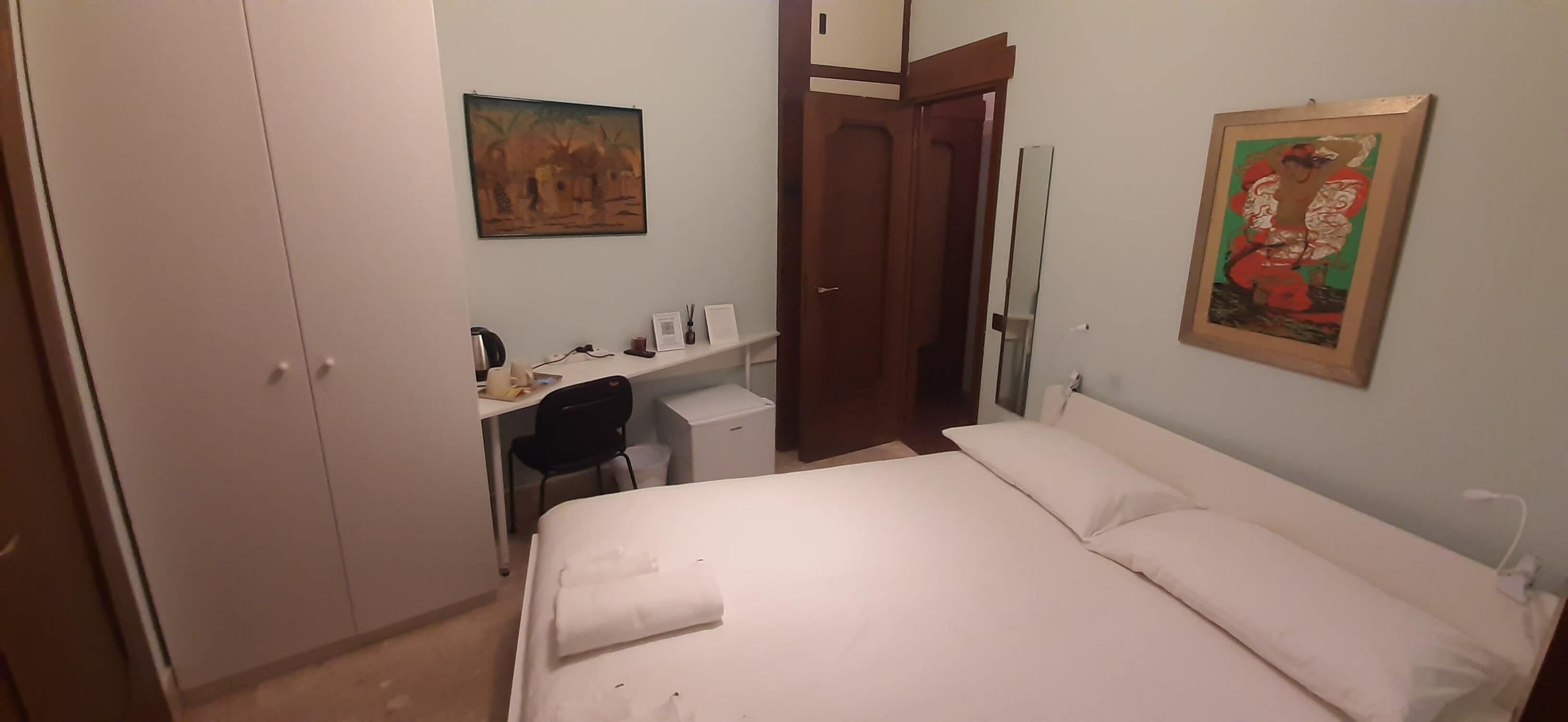 Una habitación de hotel con una cama, un escritorio con silla, un armario y dos cuadros en las paredes.