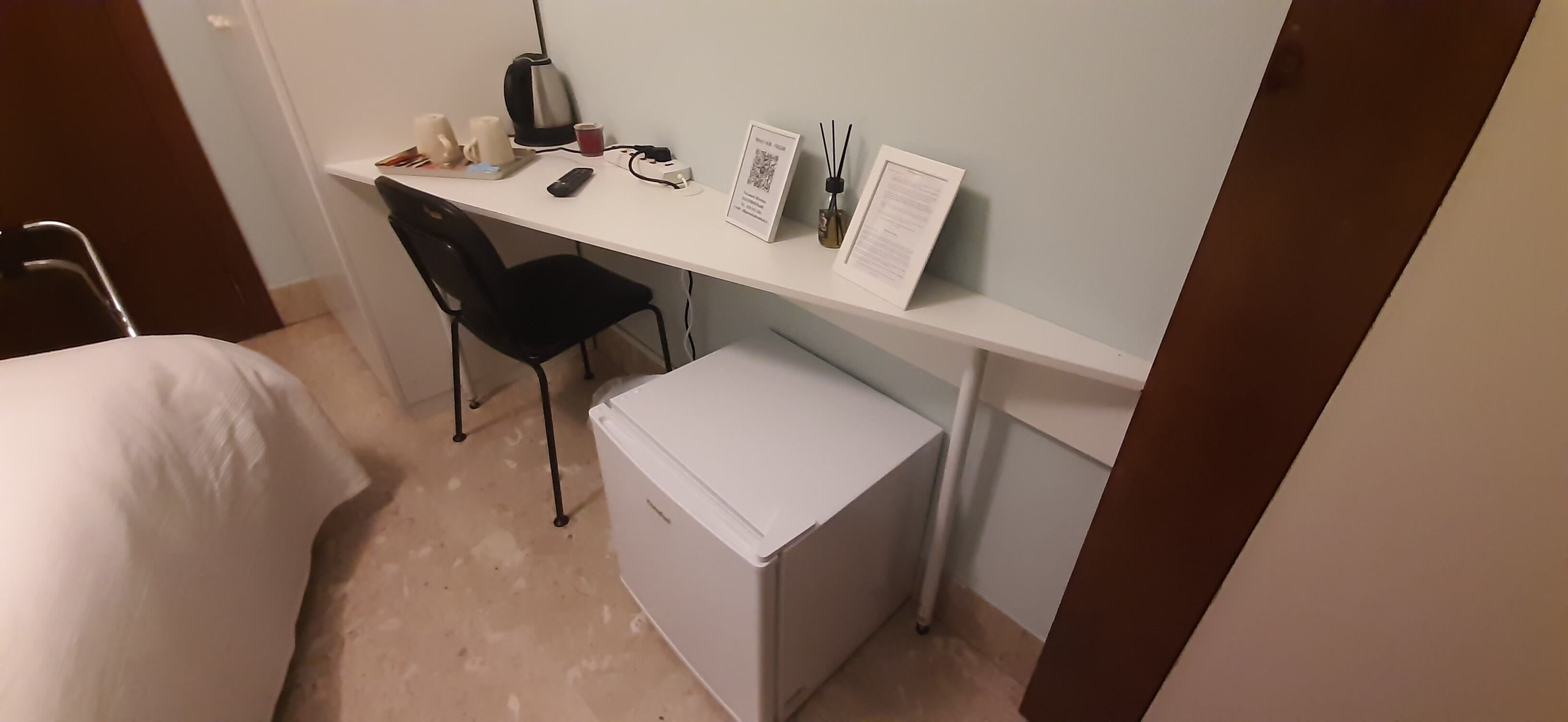 Une chambre d’hôtel compacte comprenant un lit simple, un petit bureau avec une chaise, un réfrigérateur, une bouilloire, une tasse, une télécommande et un menu encadré.