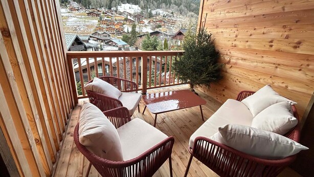 A wooden balcony with wicker furniture and a view of a snowy village. - Cosy Duplex, 3 Bedrooms + Bunk Area, Near Slopes, Garage, Ski Locker, No Smoking (Les Gets)