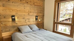 A bedroom with a wooden headboard, two bedside lamps, a bed with a grey bedspread, and a window with a view of buildings. - Cosy Duplex, 3 Bedrooms + Bunk Area, Near Slopes, Garage, Ski Locker, No Smoking (Les Gets)