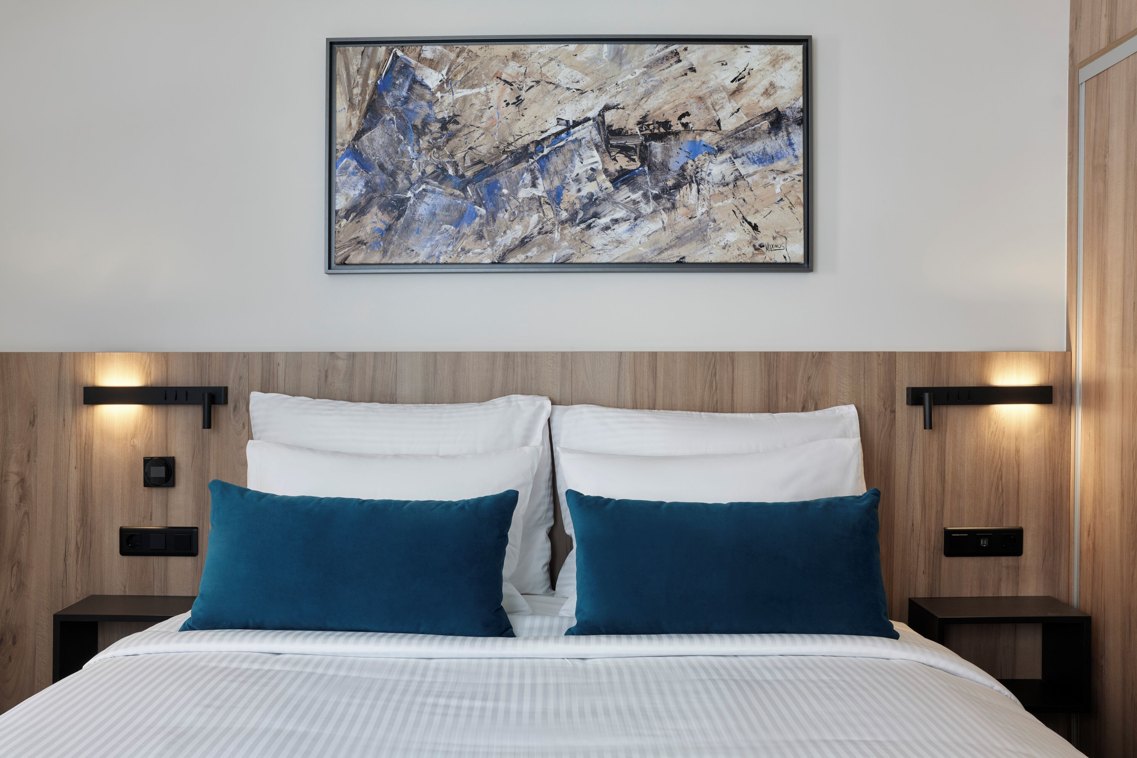 A modern bedroom with a large bed, two blue pillows, a framed abstract painting, and wooden paneling on the walls.