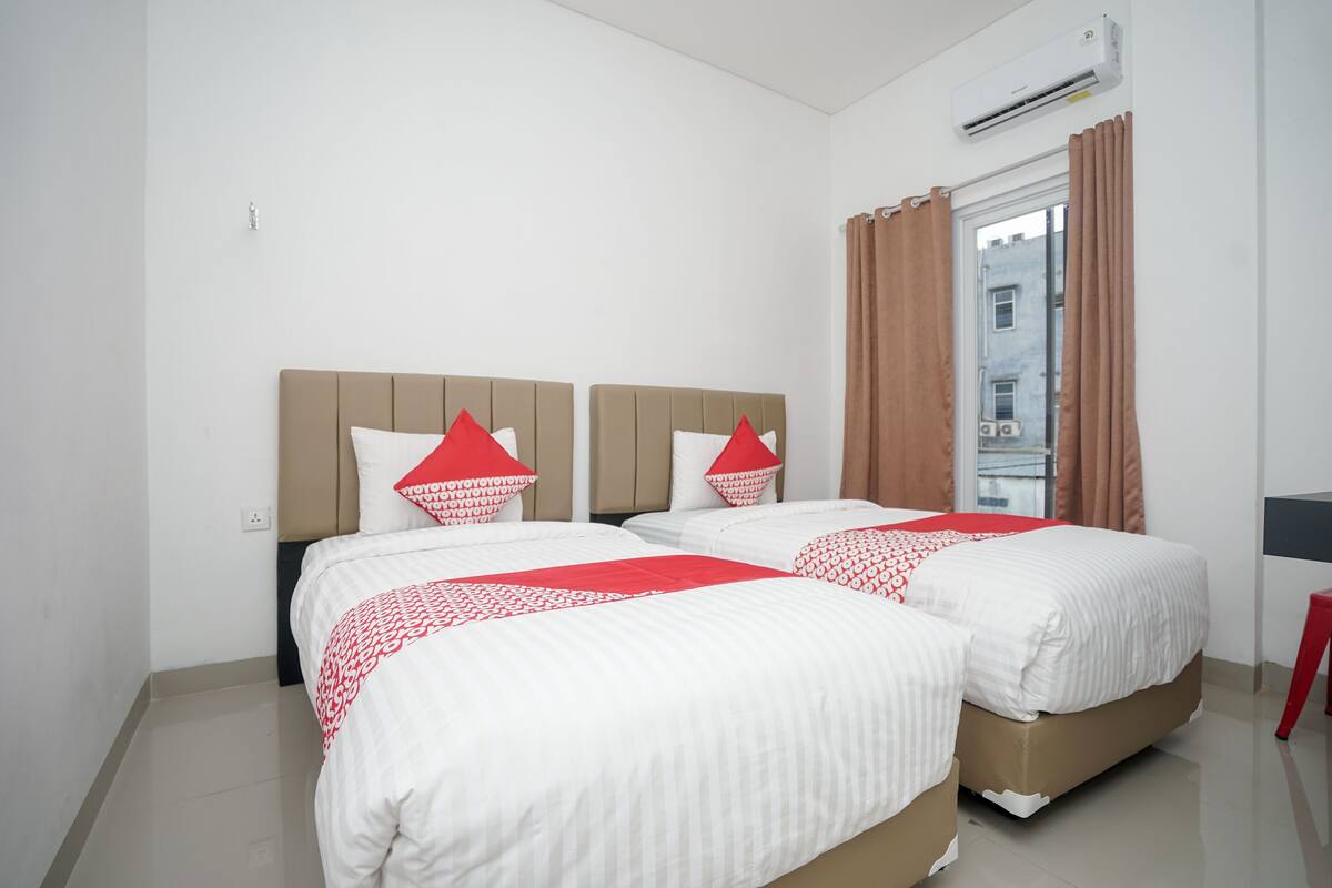 Habitación de hotel con dos camas, cada una con ropa blanca y detalles rojos, una ventana con cortinas marrones y un equipo de aire acondicionado en la pared.