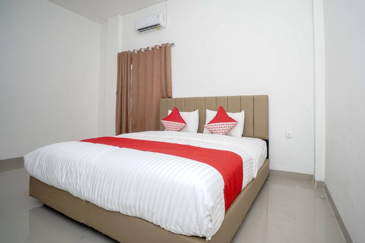 Una cama bien hecha con cabecera color beige y ropa de cama blanca, incluyendo un detalle rojo.