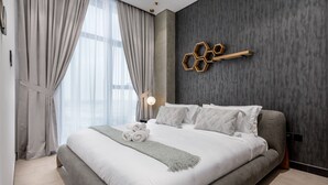 A modern bedroom with a large bed, grey bedding, a grey throw blanket, and a wall-mounted hexagonal light fixture.