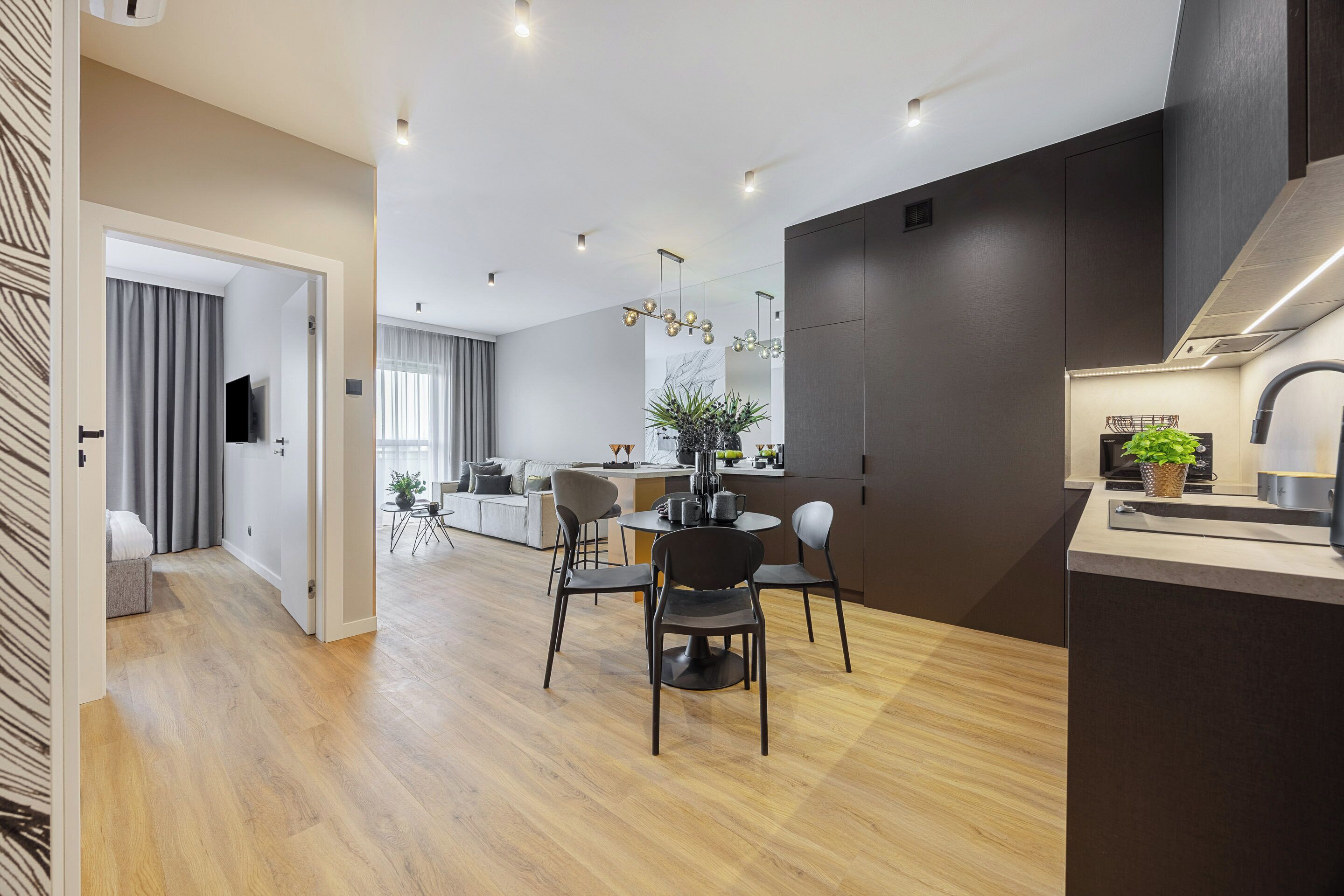 Une cuisine moderne avec des meubles de couleur sombre, un coin repas avec une table ronde et des chaises, et un salon avec un canapé et un téléviseur.