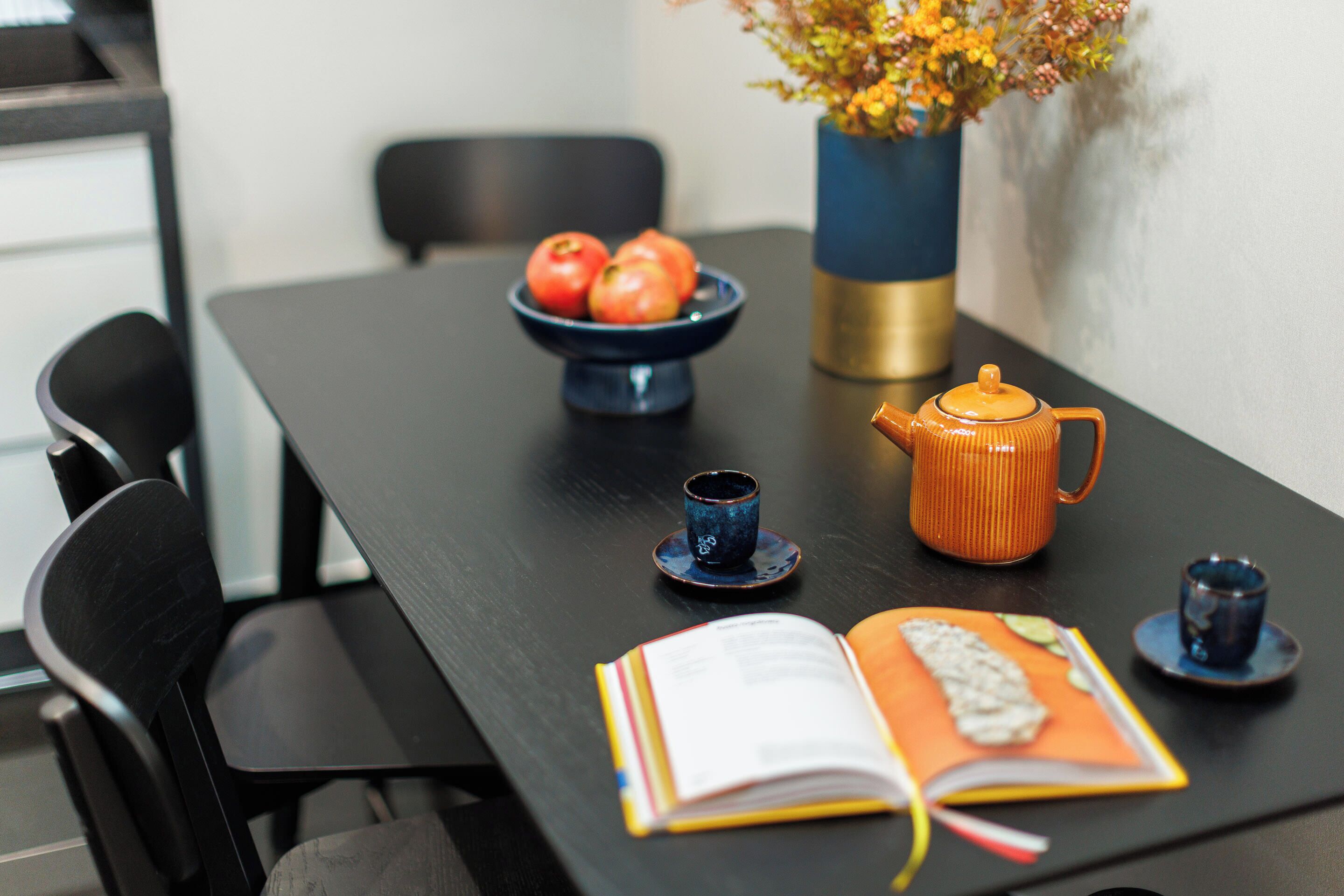 A dining table with a bowl of tomatoes, a vase with flowers, a teapot, and a book.
