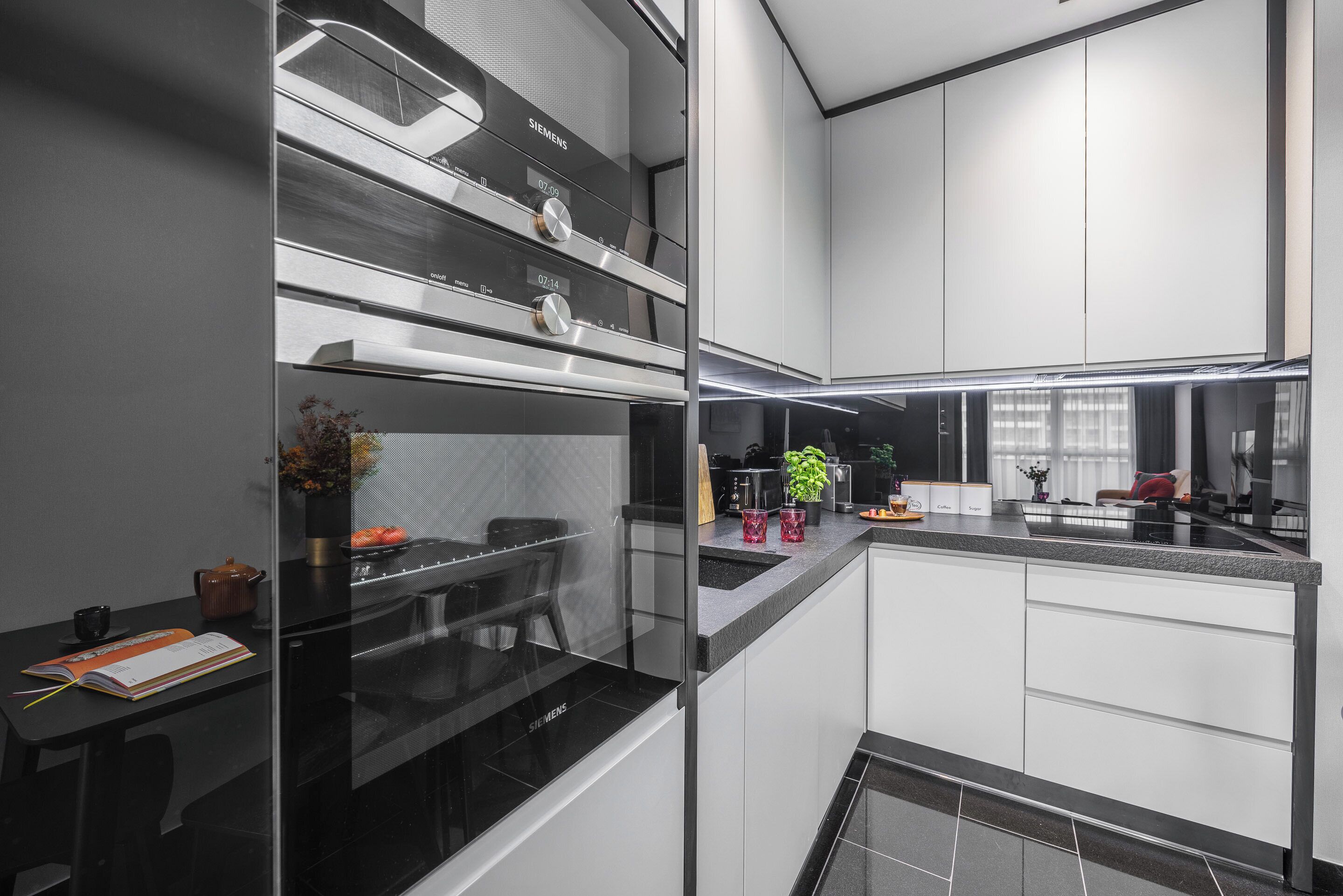 A modern kitchen with a black and white color scheme, featuring a Siemens branded oven, a glass partition, and a countertop with various items.