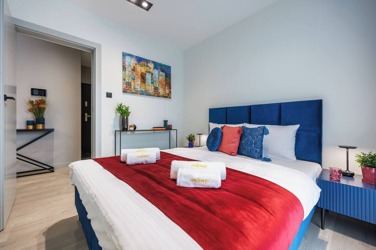 A modern hotel room with a large bed, a red throw blanket, and a blue headboard. There is a small table with a plant and a lamp.