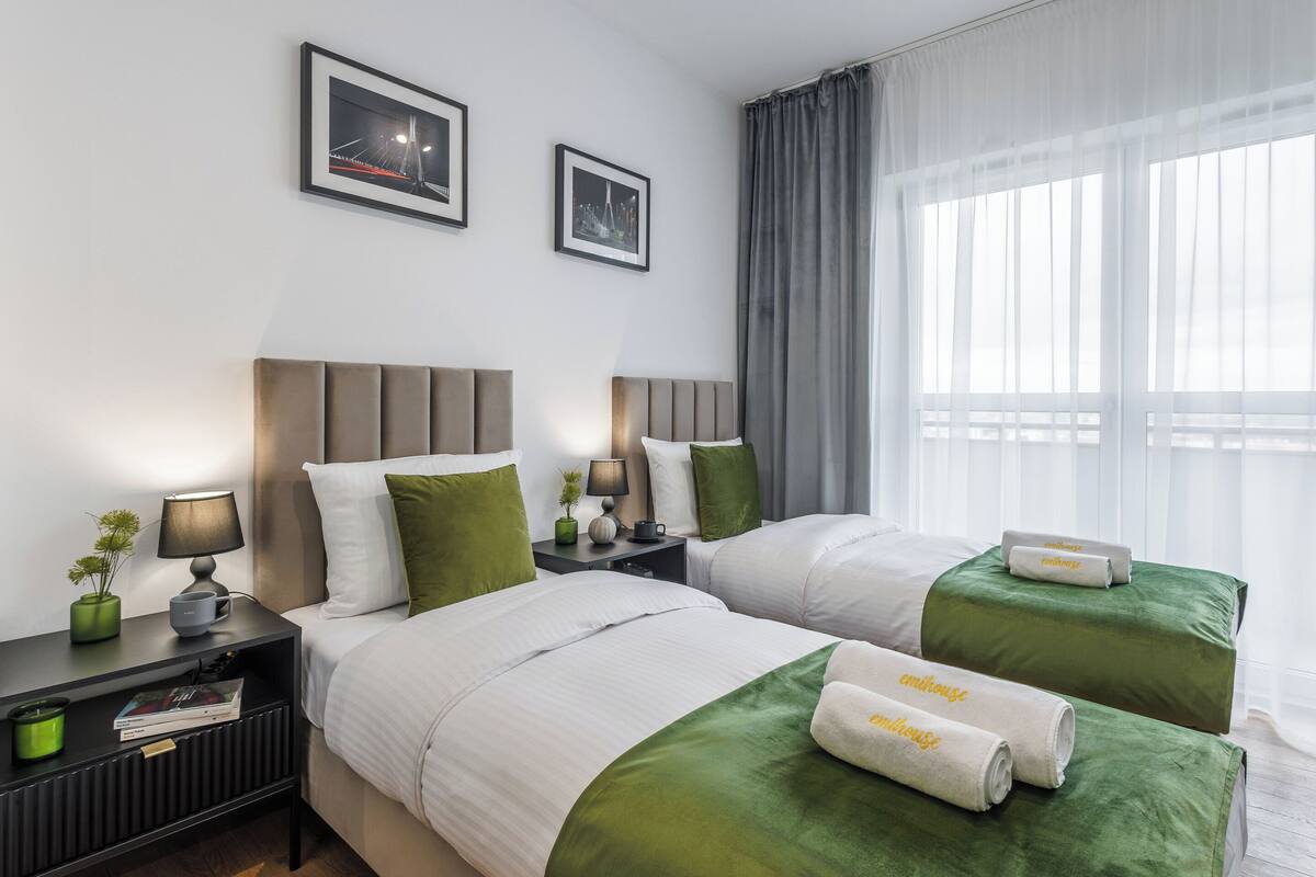 A hotel room with two beds, a nightstand with a lamp, framed pictures on the wall, and a window with curtains.