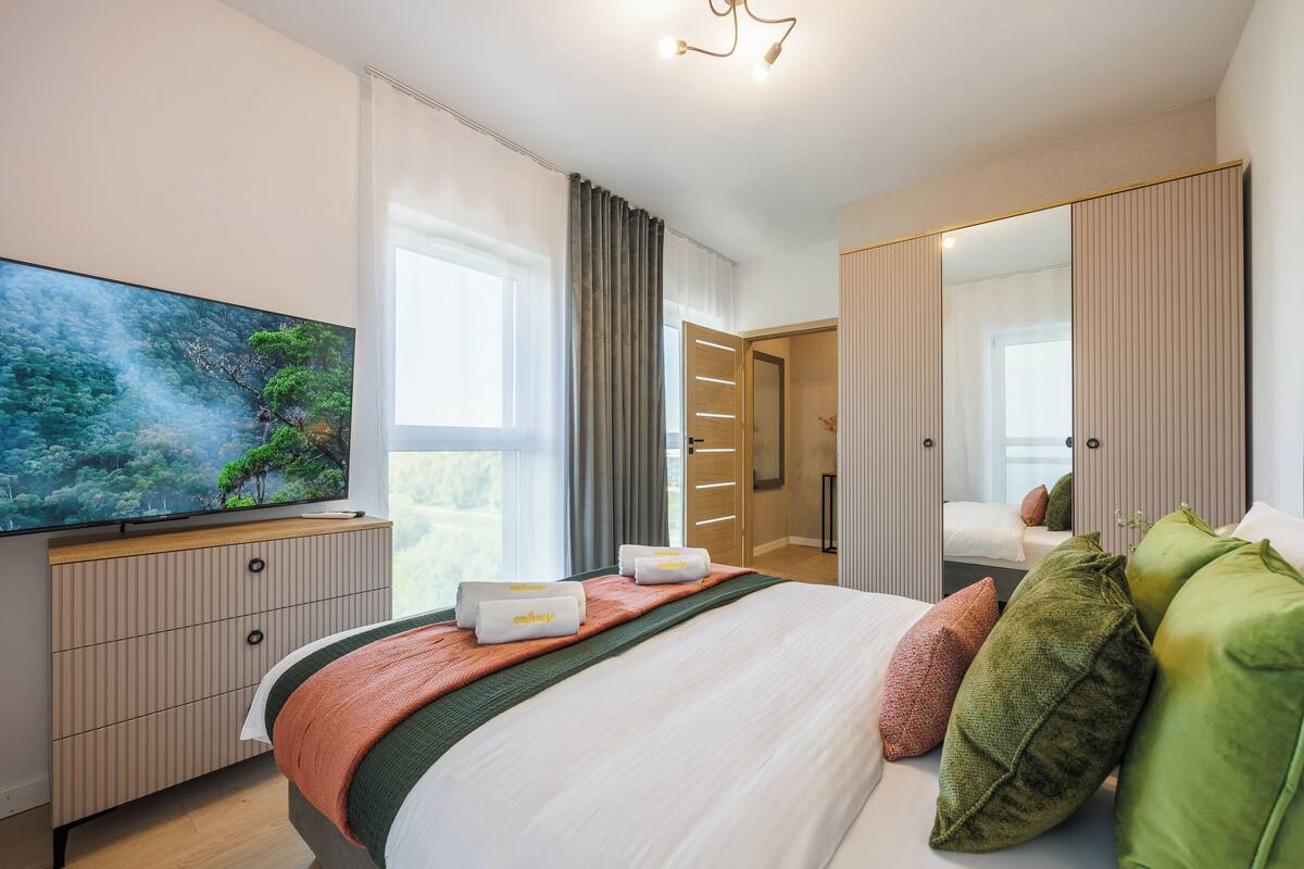 A modern hotel room with a large flat-screen TV, a bed with multiple pillows, a wardrobe, and a large window with curtains.